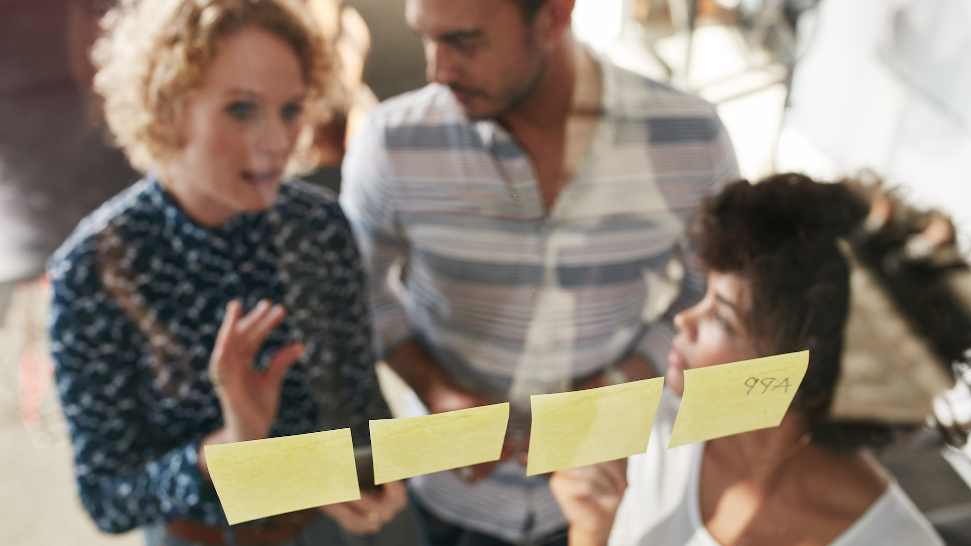 Three business people having a meeting in office. They are standing in front of glass wall with post it notes and discussing.