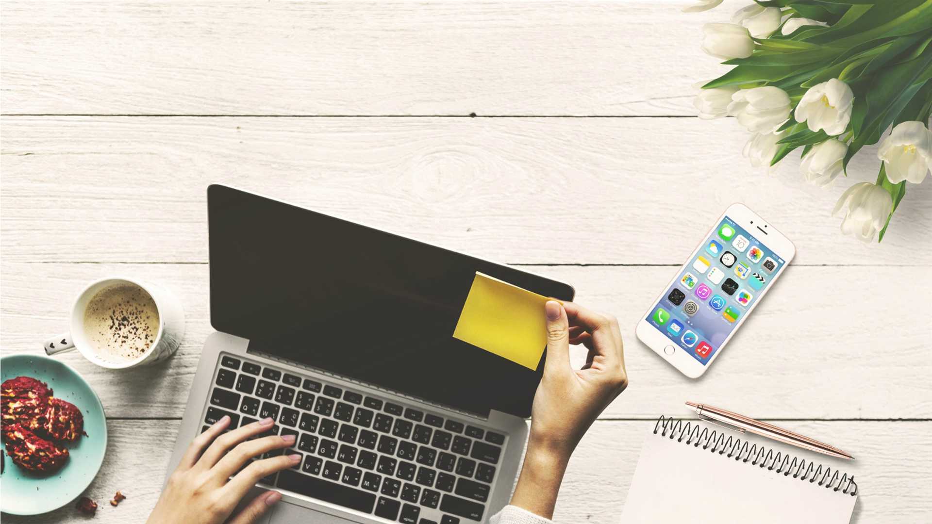person working on a laptop with a sticky note, phone, flowers and breakfast