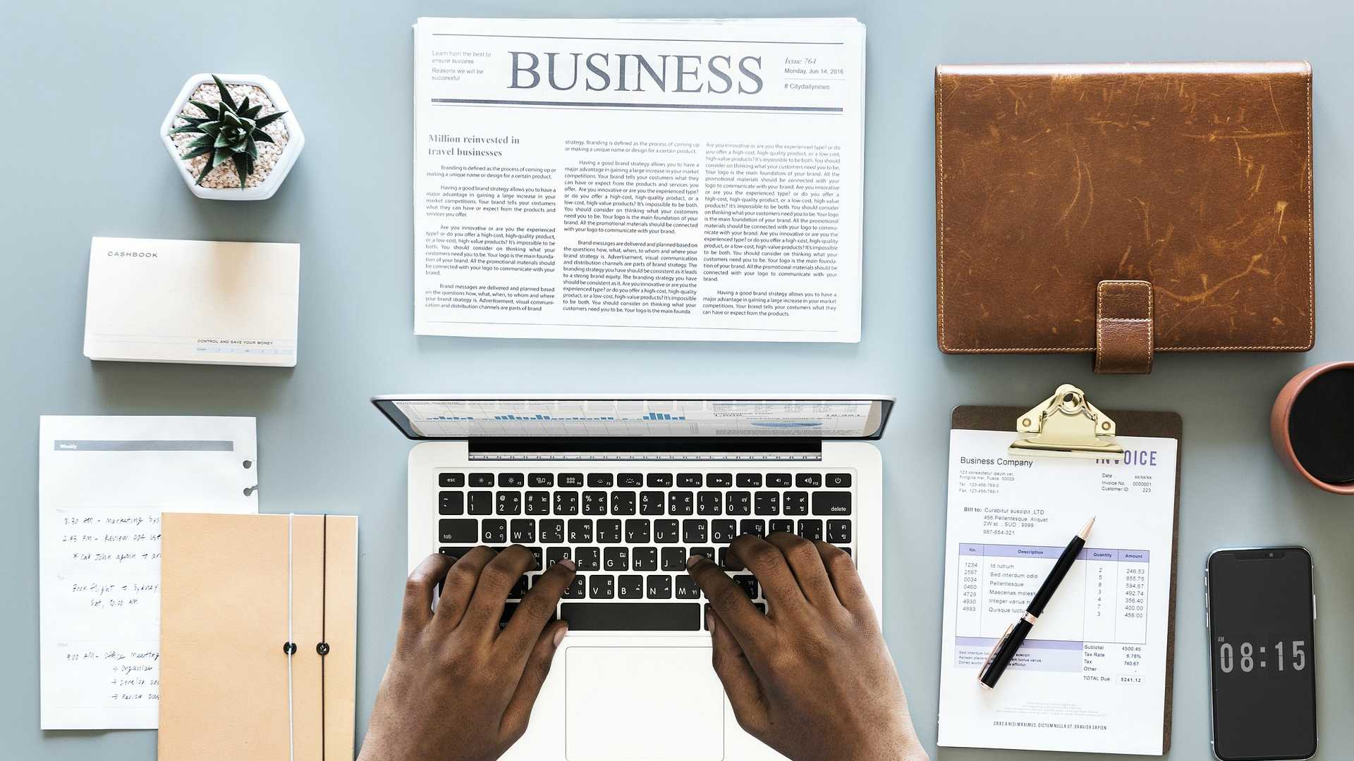 man working on a laptop with a business newspaper and other materials