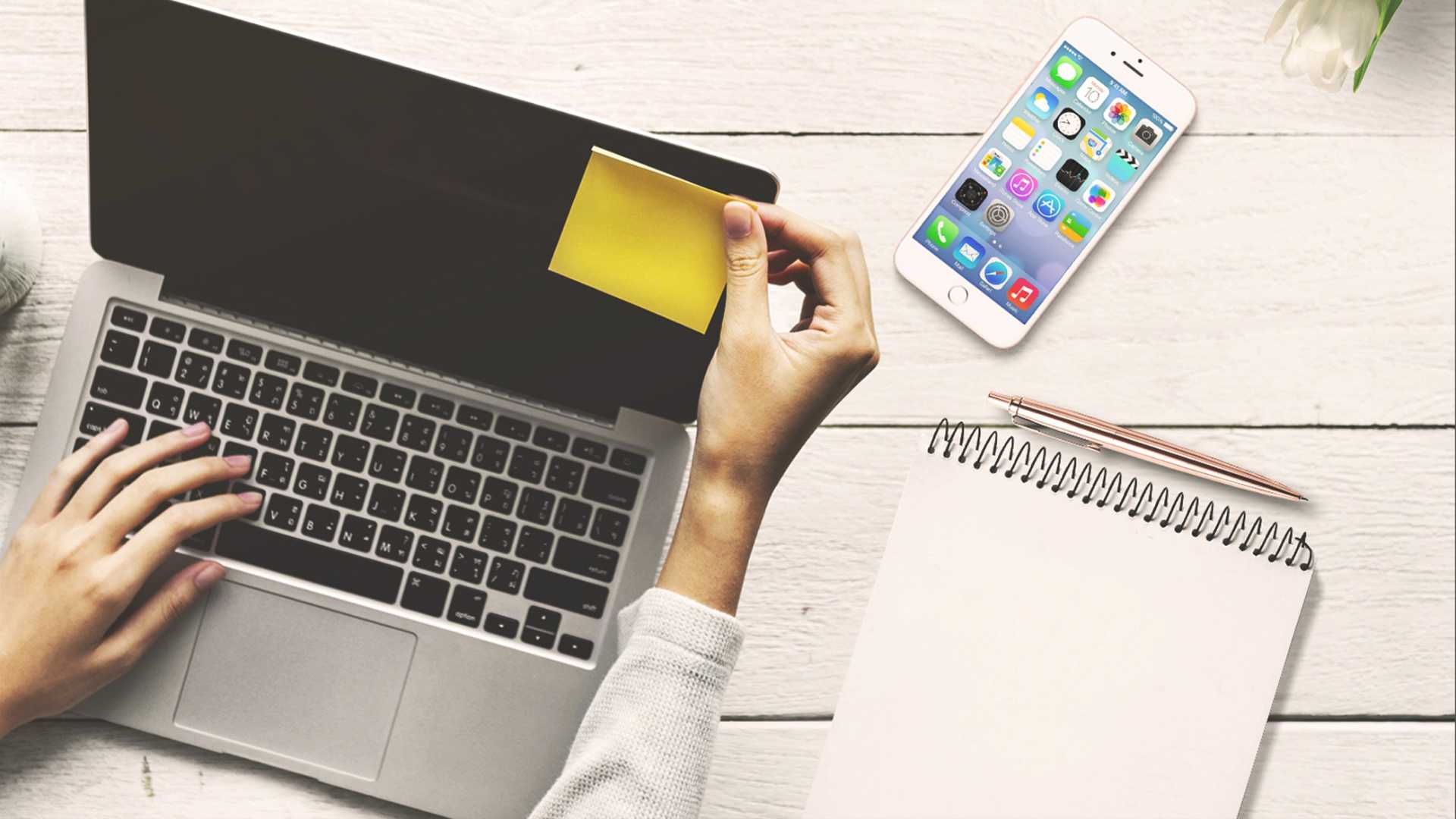 woman putting a sticky note on her laptop; phone and notepad on her desk