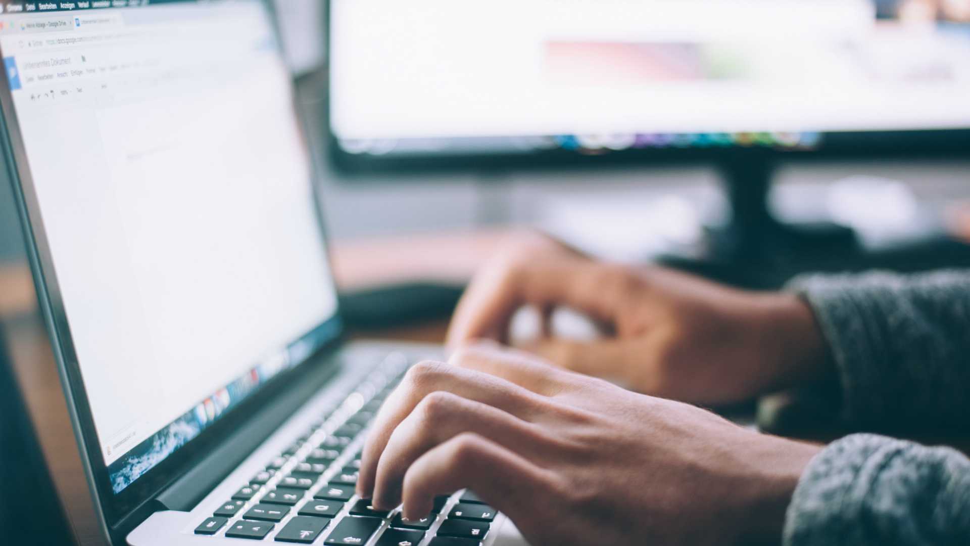 person typing on a laptop with a larger monitor on the desk