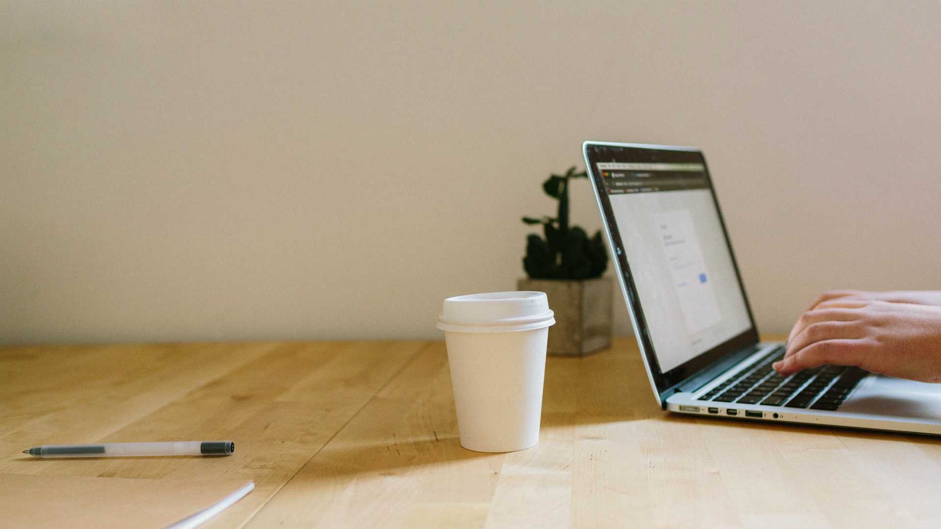 person working on a laptop at a table with a plant, a pen and a coffee cup
