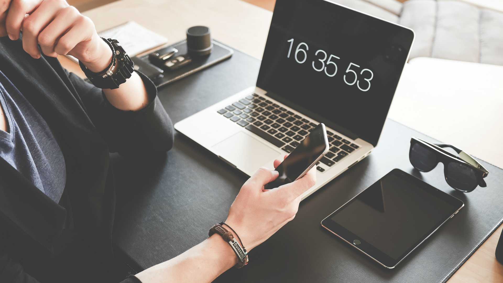 person using a mobile phone at a desk with a laptop that has the time on it