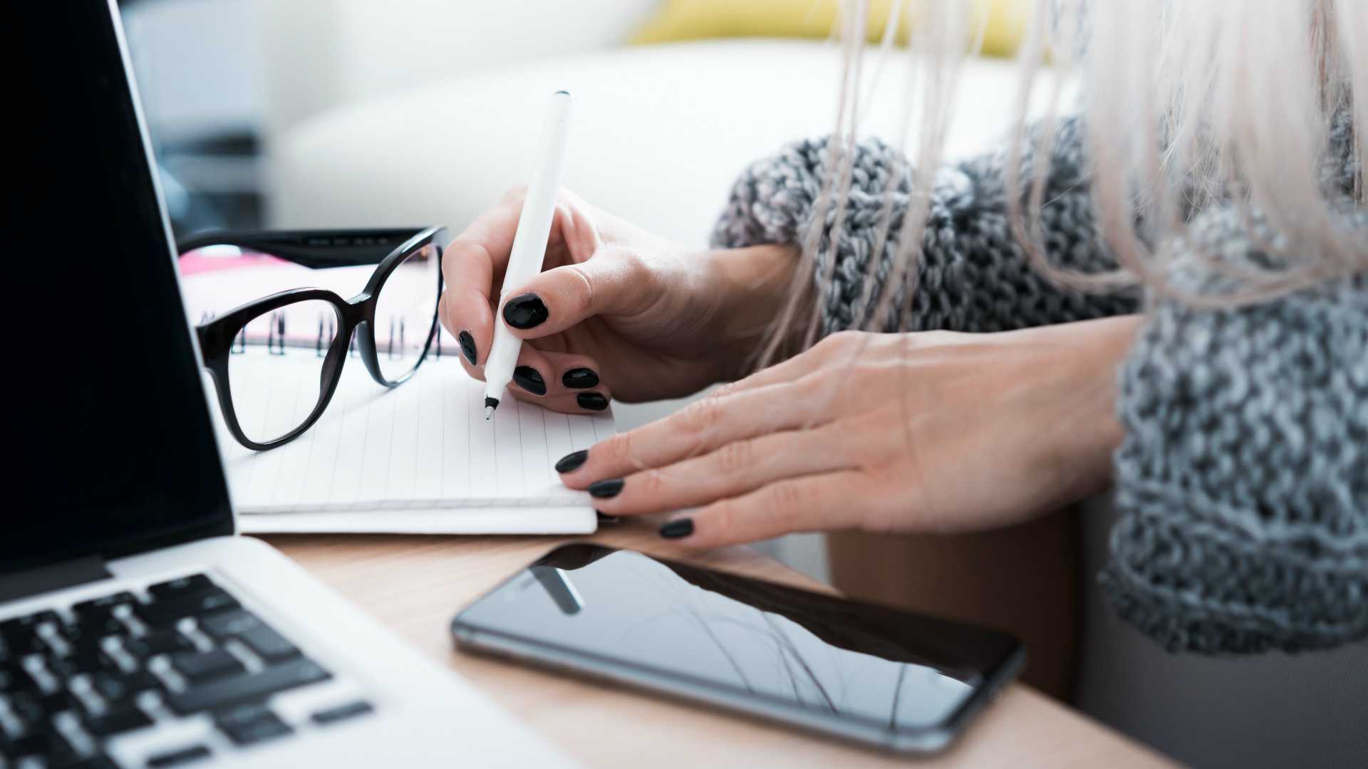 woman taking notes at her desk with a laptop and smartphone