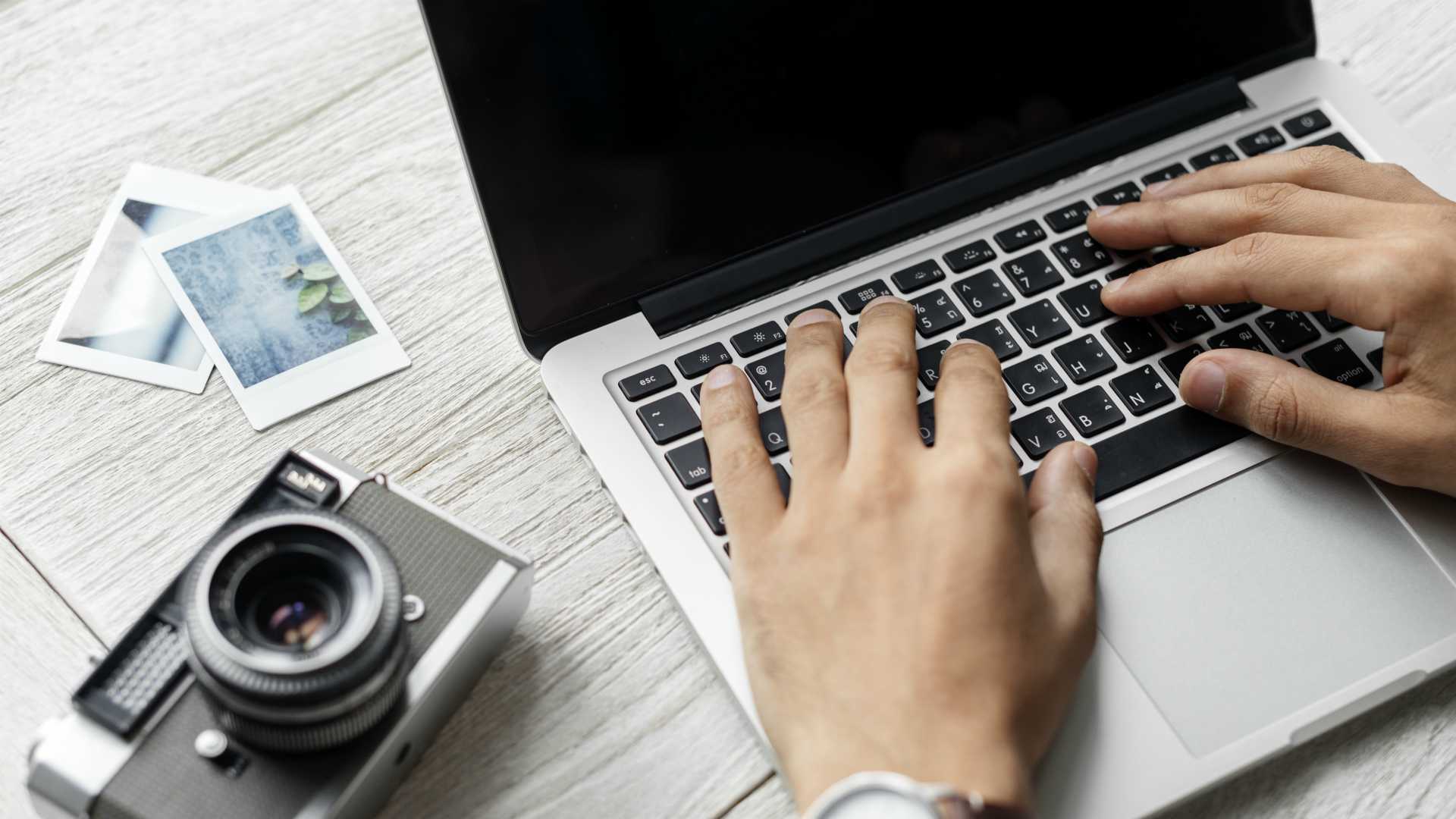 person typing on a laptop with photos and a camera on the table