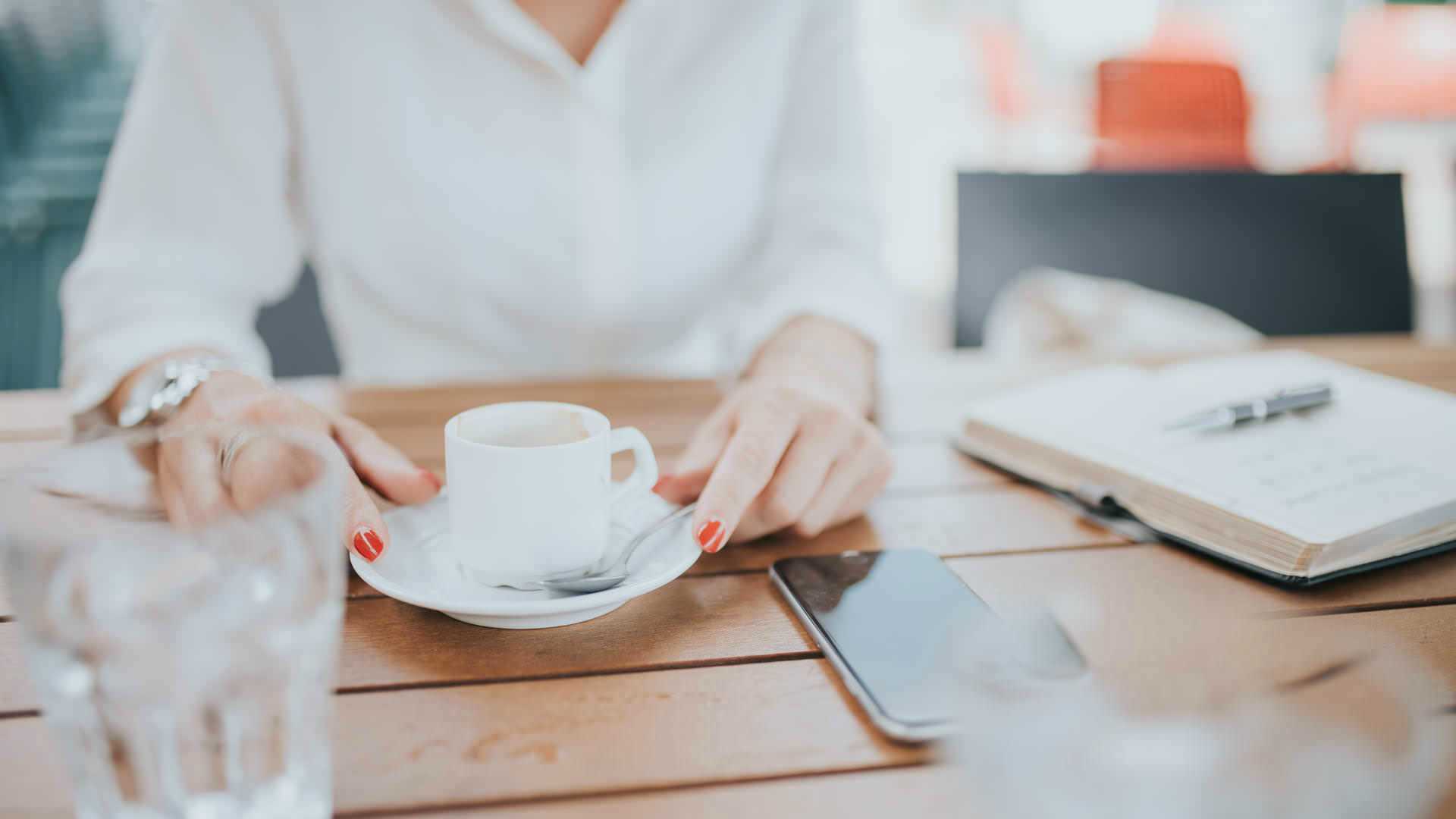 woman sitting at a table with a smartphone, an open notebook with a pen, and a glass of water; positioning her teacup and waiting to talk