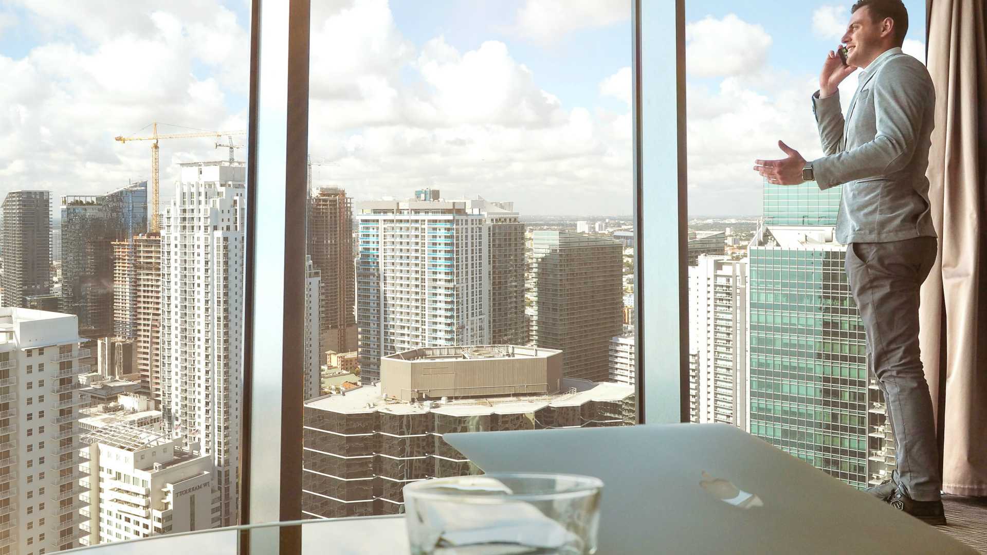 businessman talking on his cell phone in his office with floor-to-ceiling walls overlooking the city