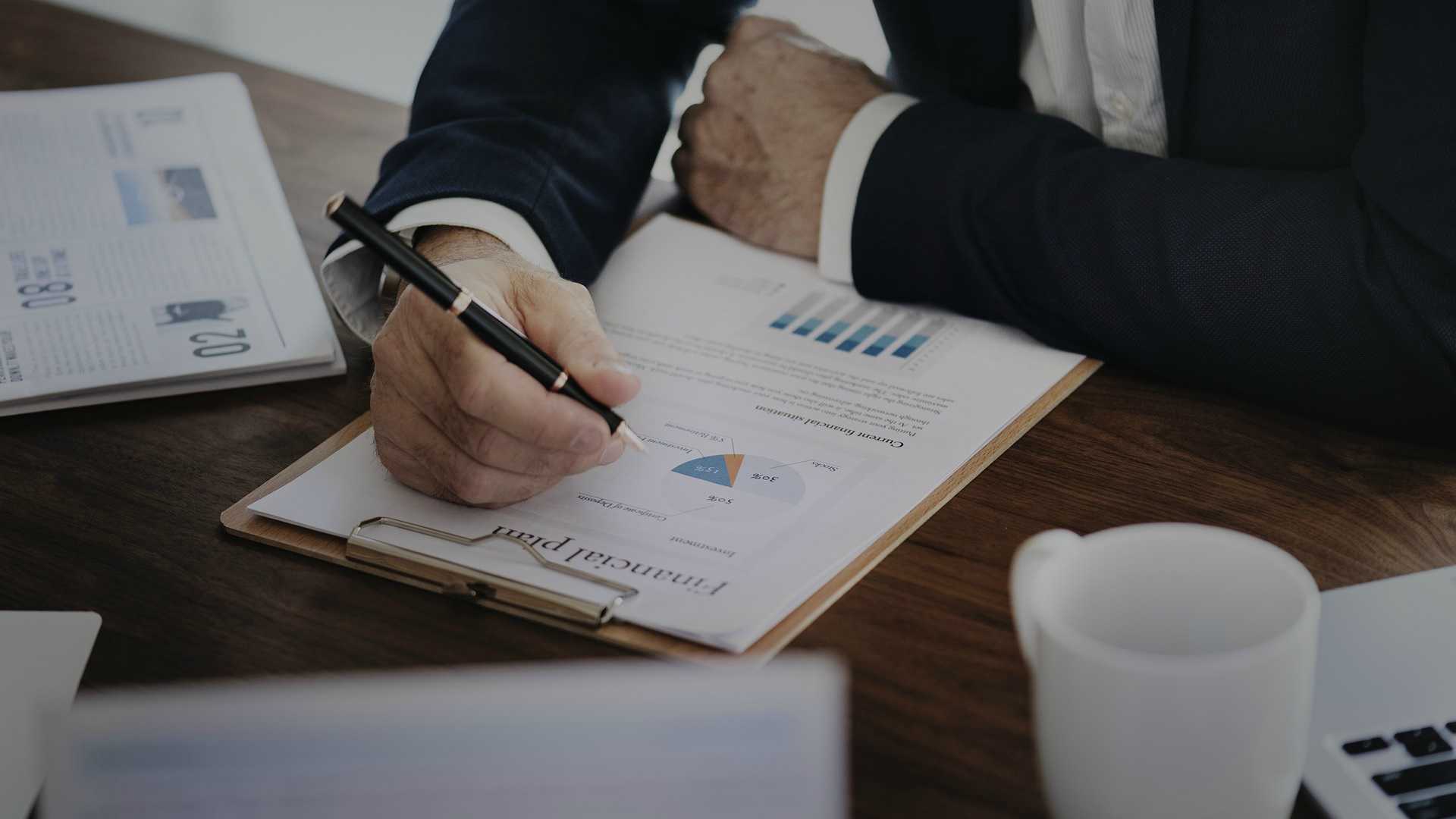 businessman looking at a piece of paper with charts on it and taking notes; on his desk is other papers with charts and a coffee mug