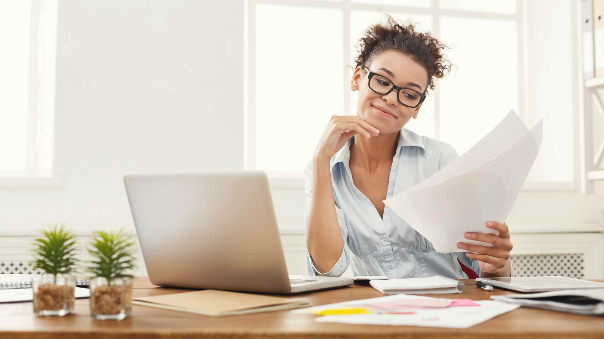 happy young employee looking at papers in her hand and sitting at a desk with a laptop, two small plants and other papers