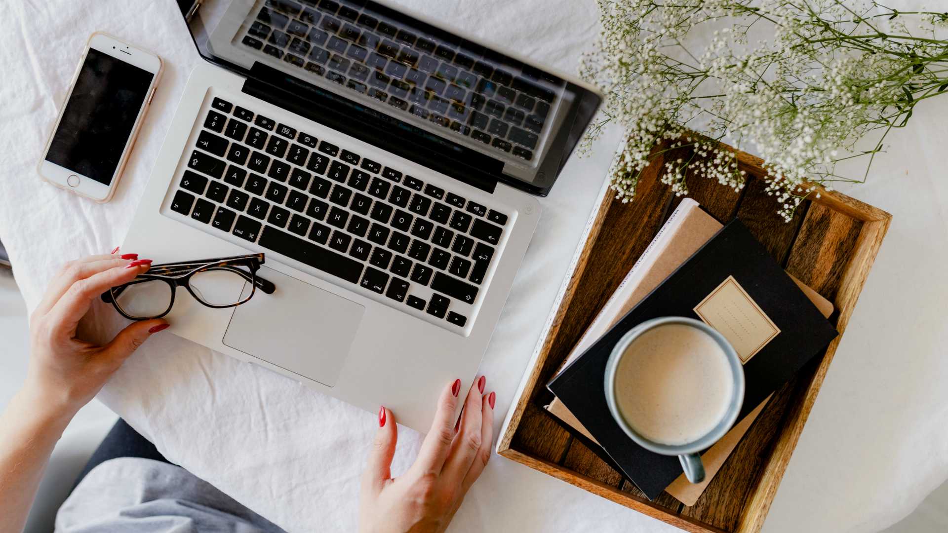 woman using a laptop and holding glasses; a plant, smartphone, books/notebooks and coffee are on her desk