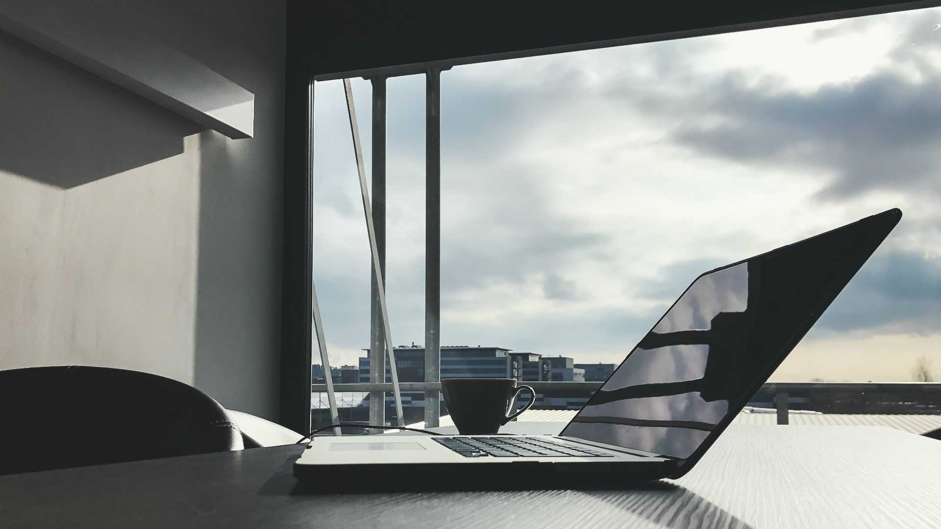 office with a large window; laptop and cup of coffee on the desk