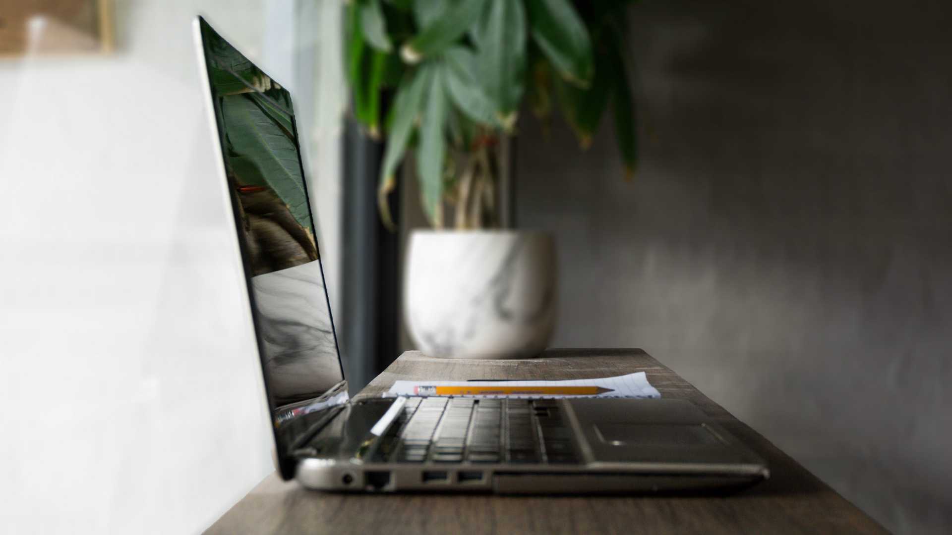 side view of a desk in front of a window with a laptop, notepad, pencil and plant on it