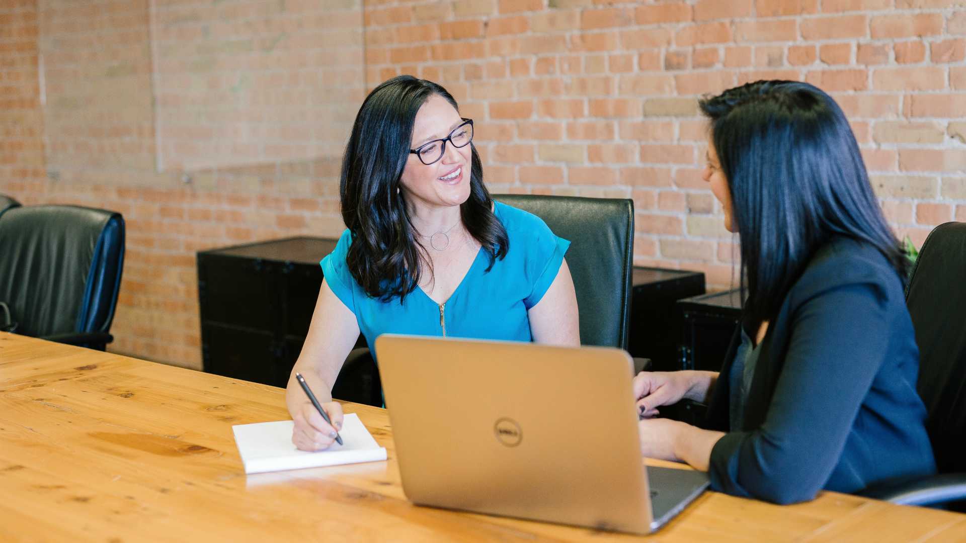 two women meeting in a conference room; one is using a laptop, and one is taking notes on a pad of paper