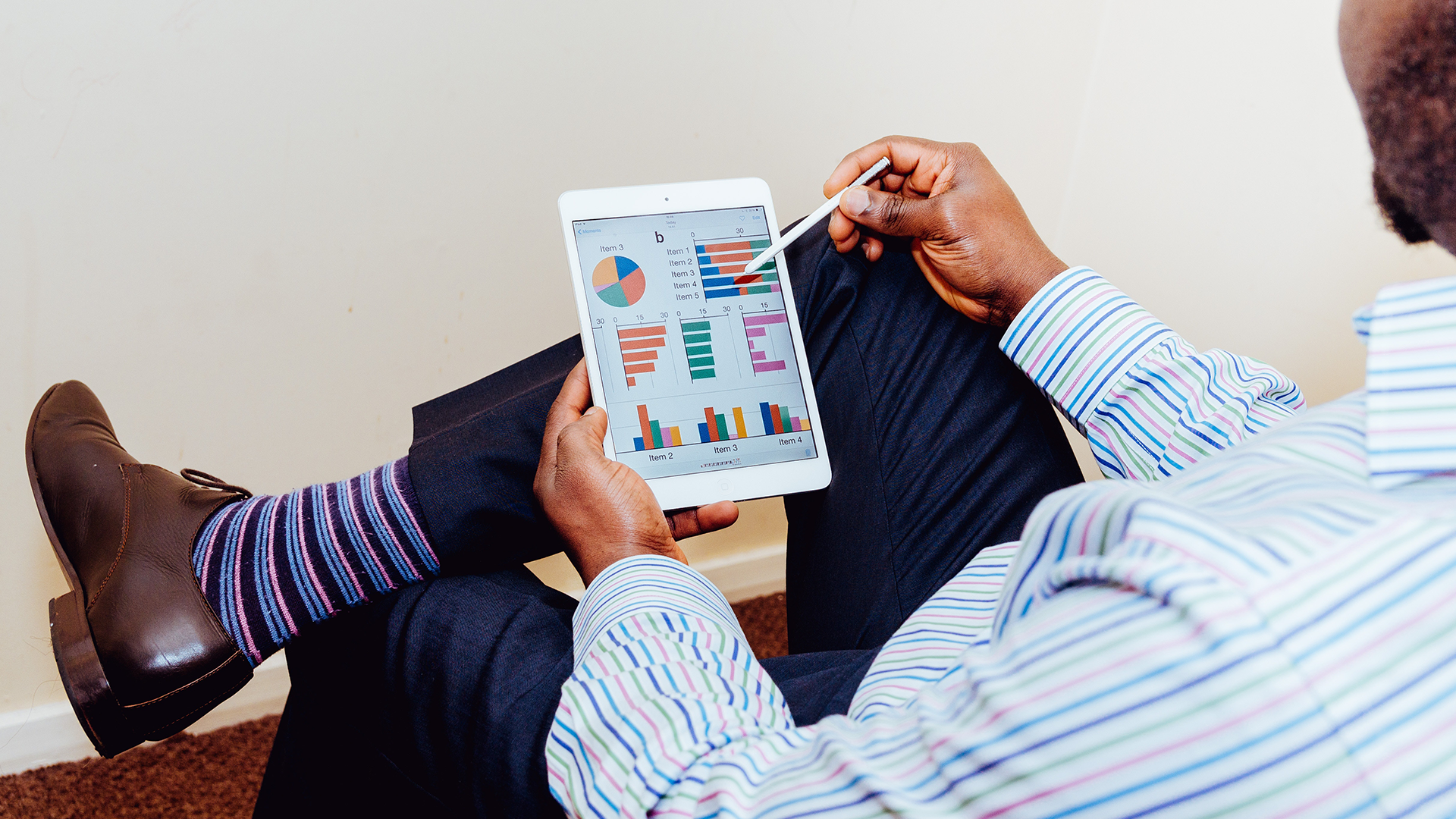 man looking at graphs and charts on a tablet, leaning on his crossed legs; using a stylus to point to a chart