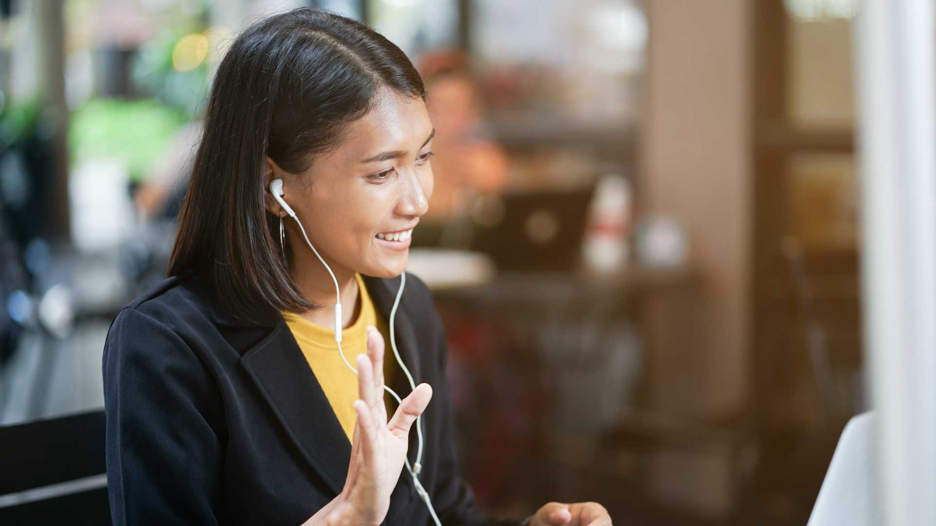 businesswoman wearing earbuds waving at someone she sees on a laptop