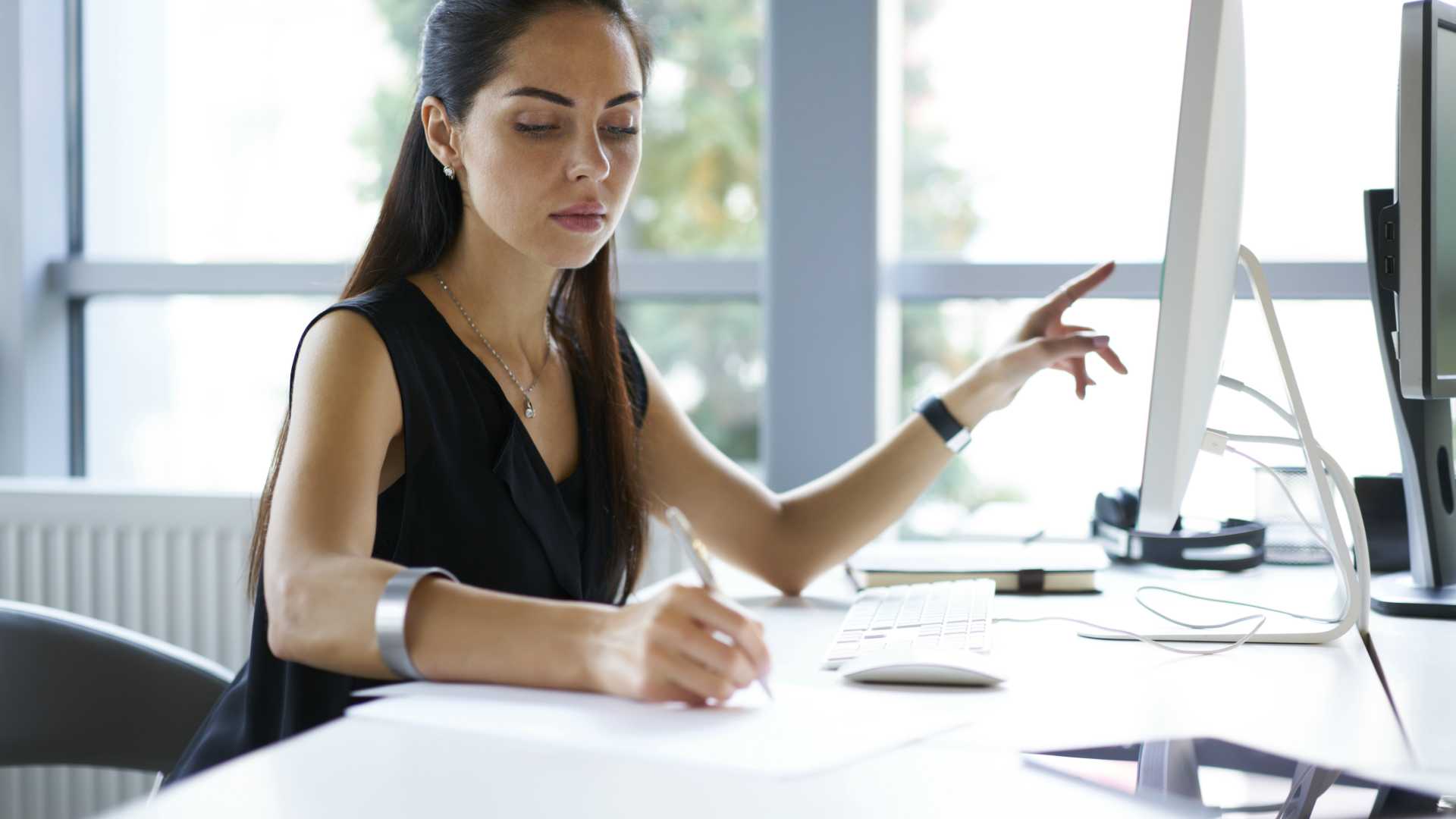businesswoman copying something in her notes that she's point to on her desktop