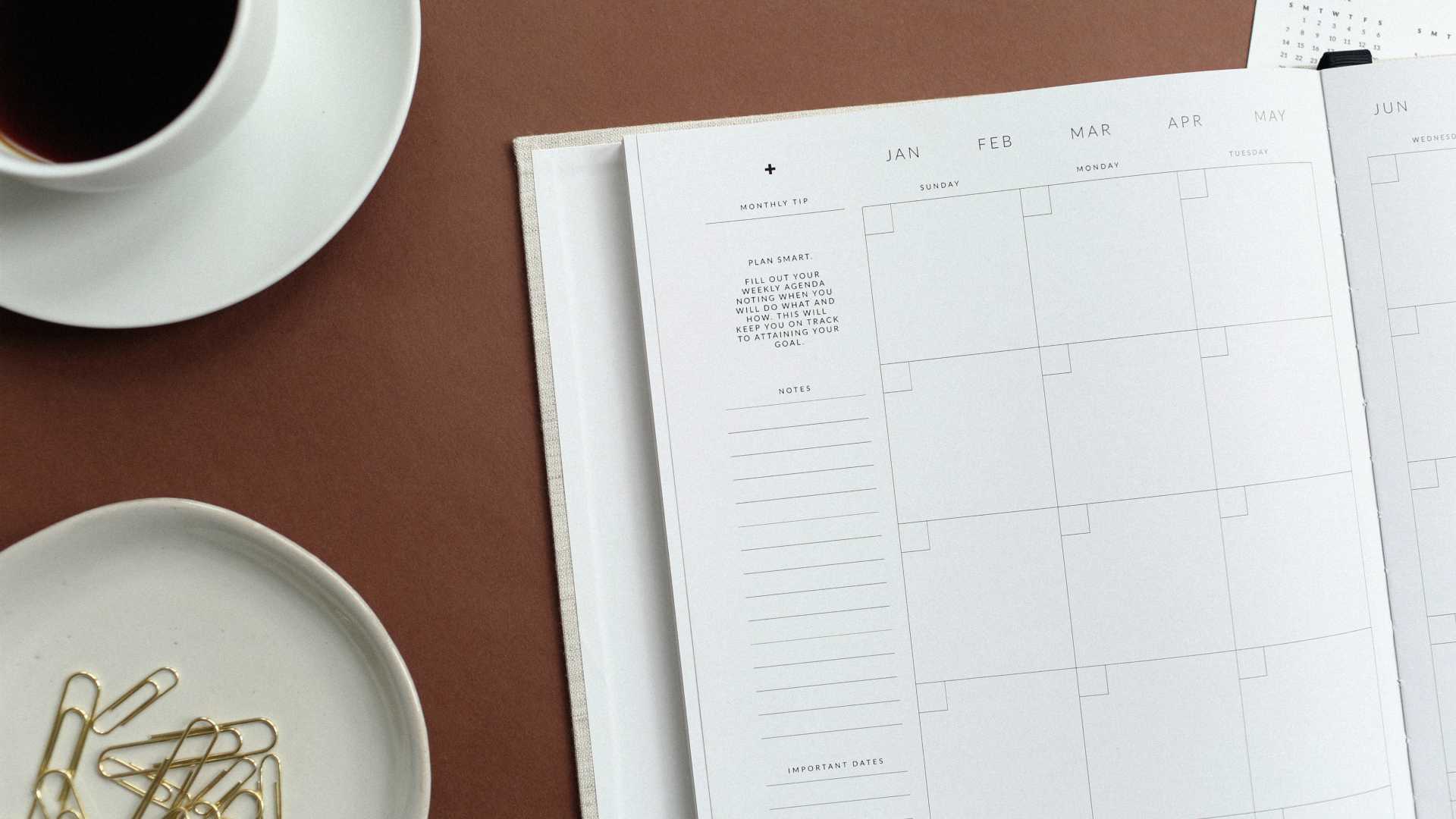 open planner on a desk with a plate of paper clips and a cup of coffee