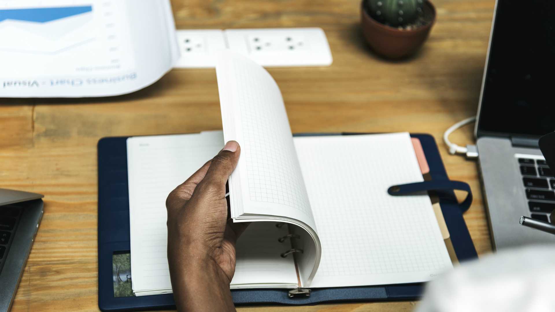 woman's hands flipping through a notebook; sitting at a table with laptops and business charts