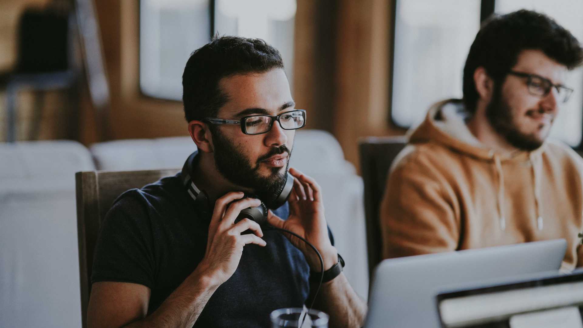 man putting on headphones and looking at his computer monitor; another man works next to him