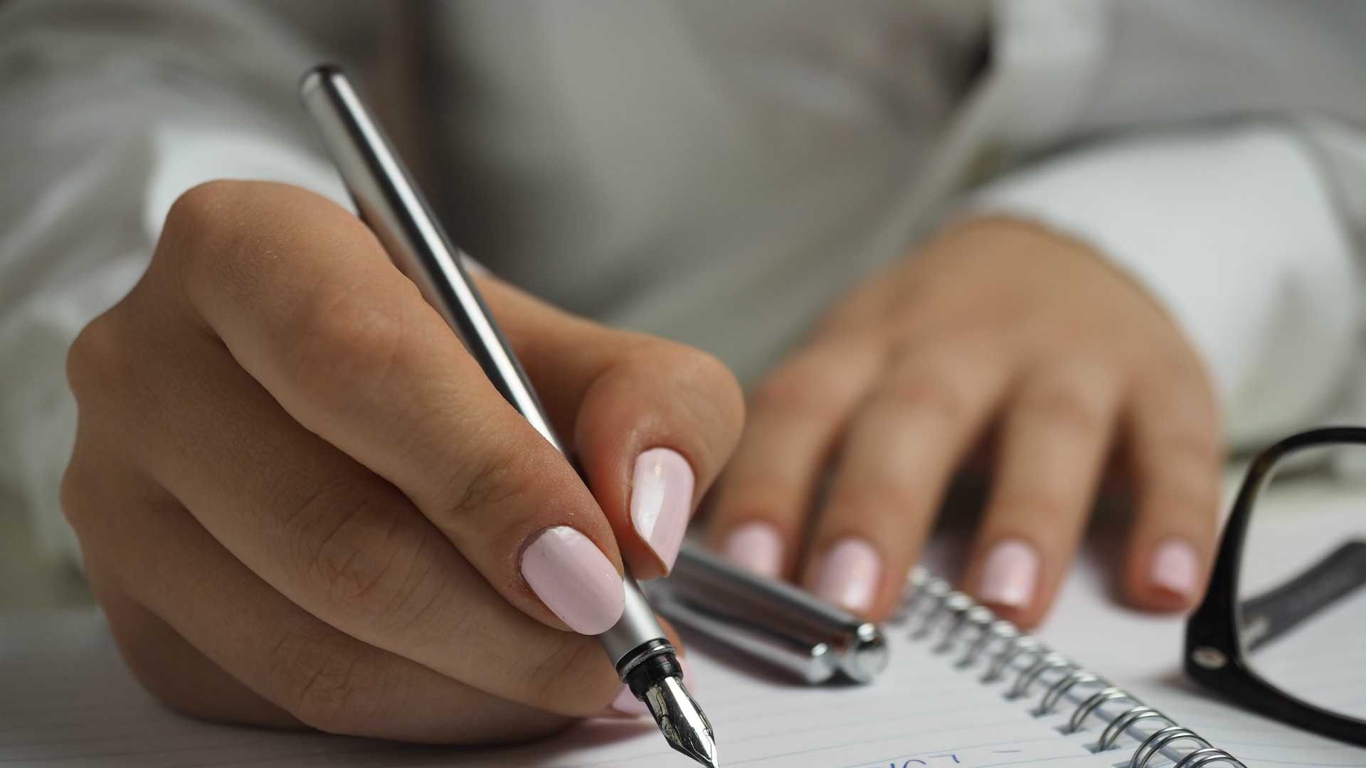 woman's hands writing in a notebook; glasses are sitting on the notebook
