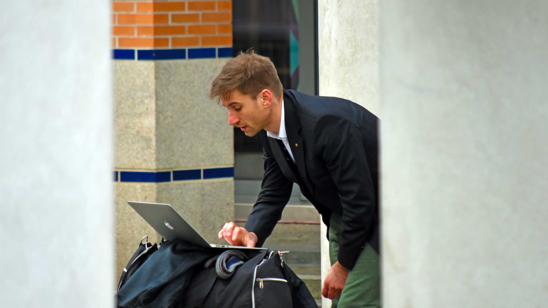man in suit checking his laptop outside, propped up on his duffel bag