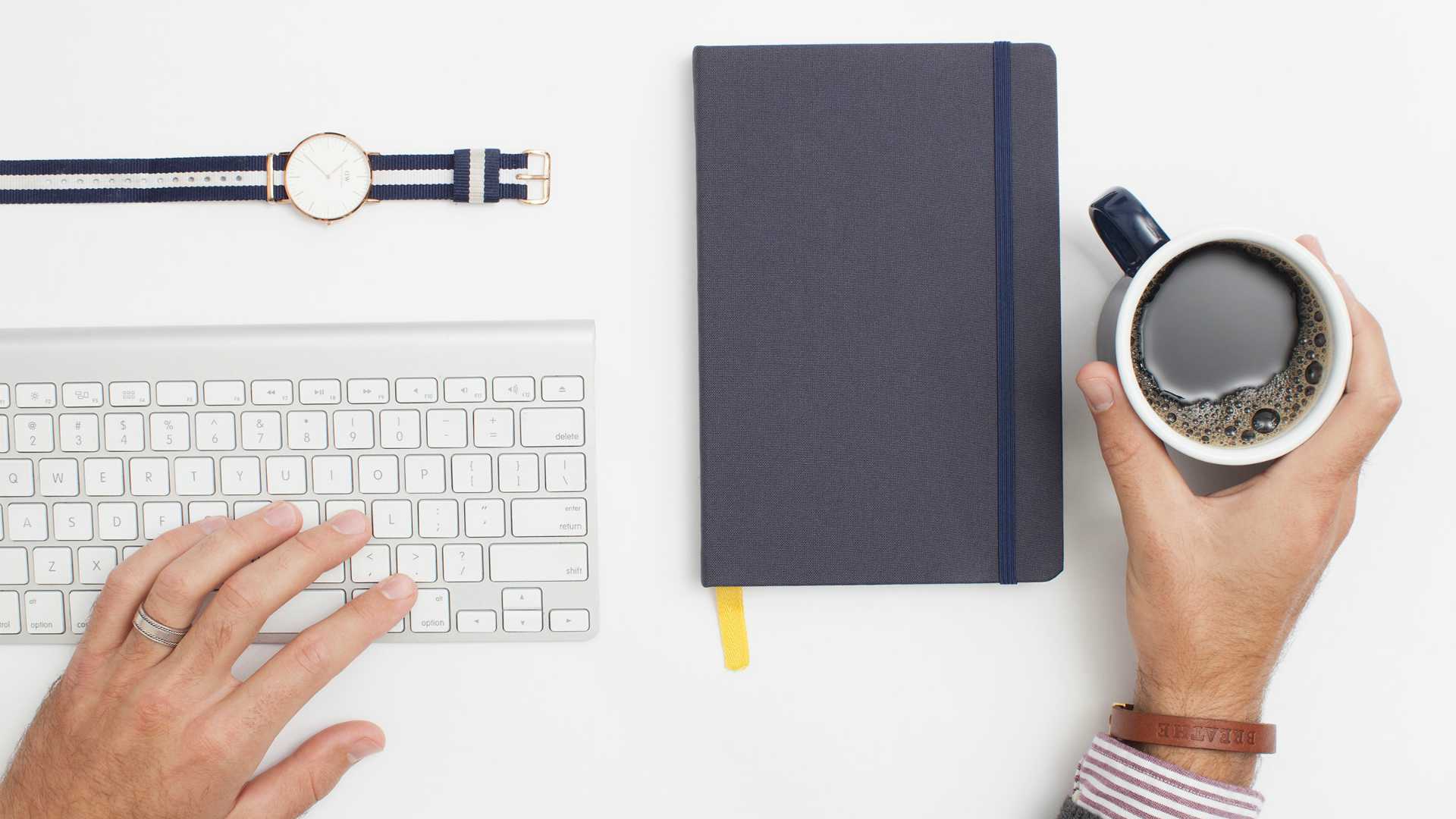 man's hands typing on a keyboard and picking up a cup of coffee; a watch and a journal are on the desk