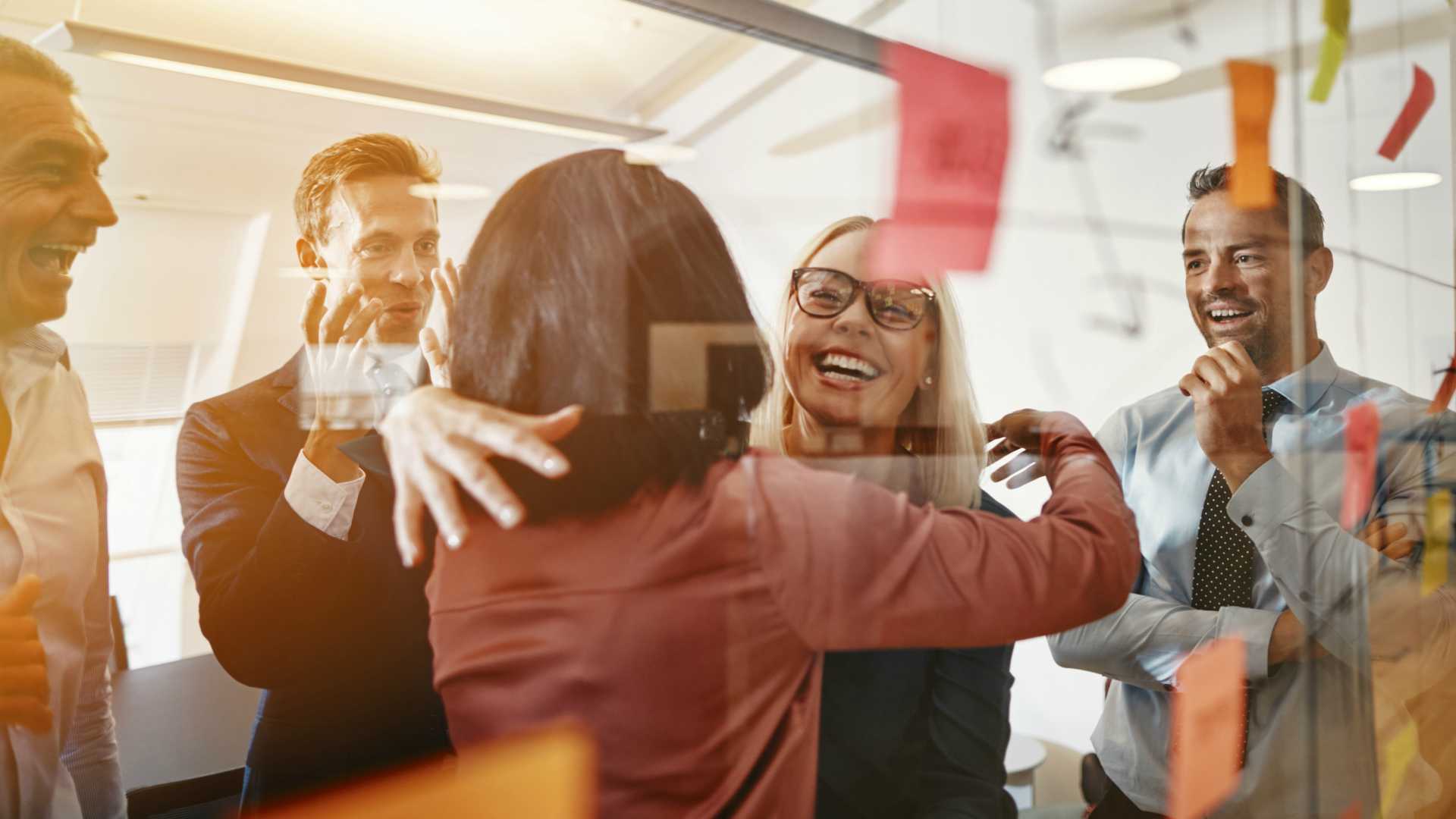 Two smiling young businesswomen hugging while brainstorming with sticky notes on a glass wall with colleagues in a modern office