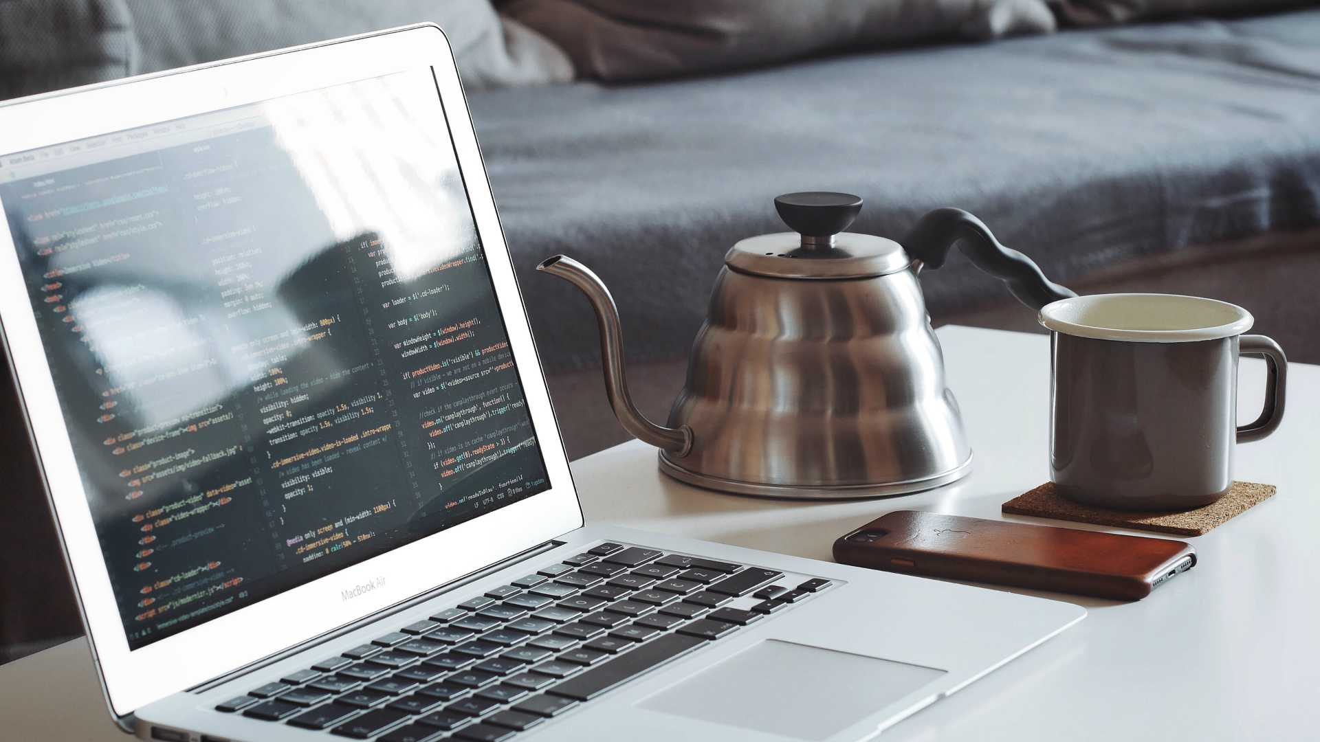 laptop with code on the screen, on a coffee table with a coffee/teapot and a mug; couch in the background