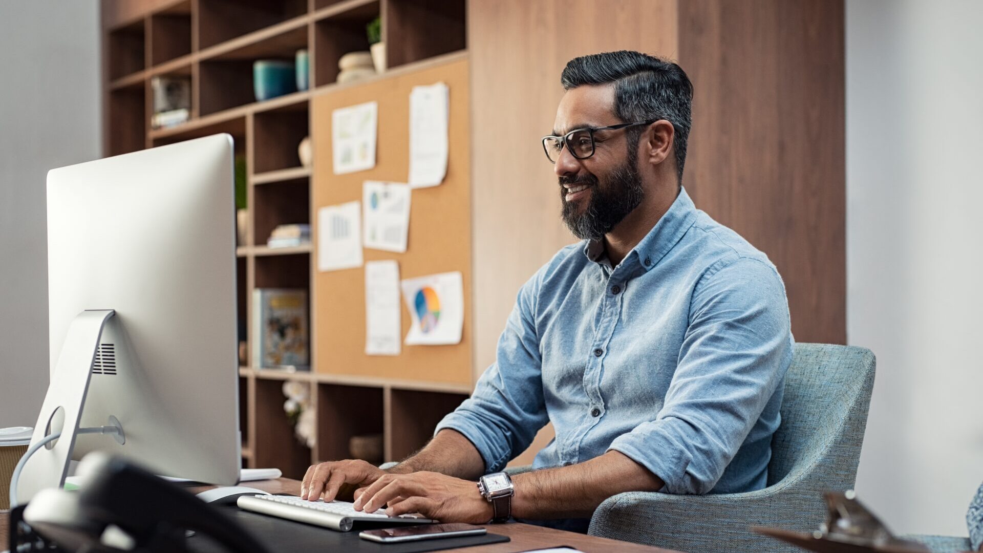 Smiling creative business man typing on desktop computer in office. Mature middle eastern businessman working at modern office space. Happy hispanic man wearing casual and eyeglasses working on his computer