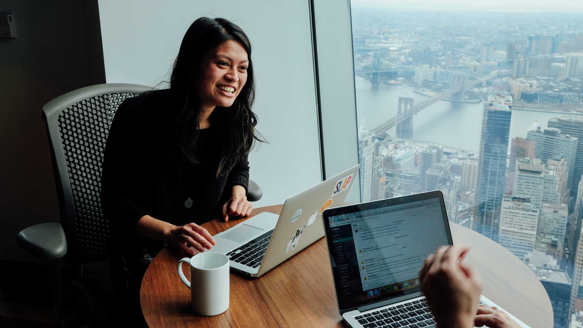 Businesswoman sitting across from colleague in front of a window in high-rise office, discussing business strategy