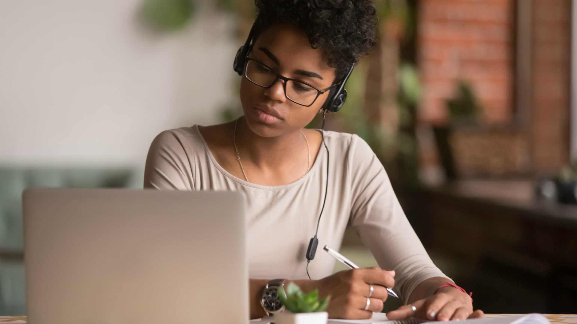 Focused mixed race woman wearing headphones watching webinar write notes study online with skype teacher