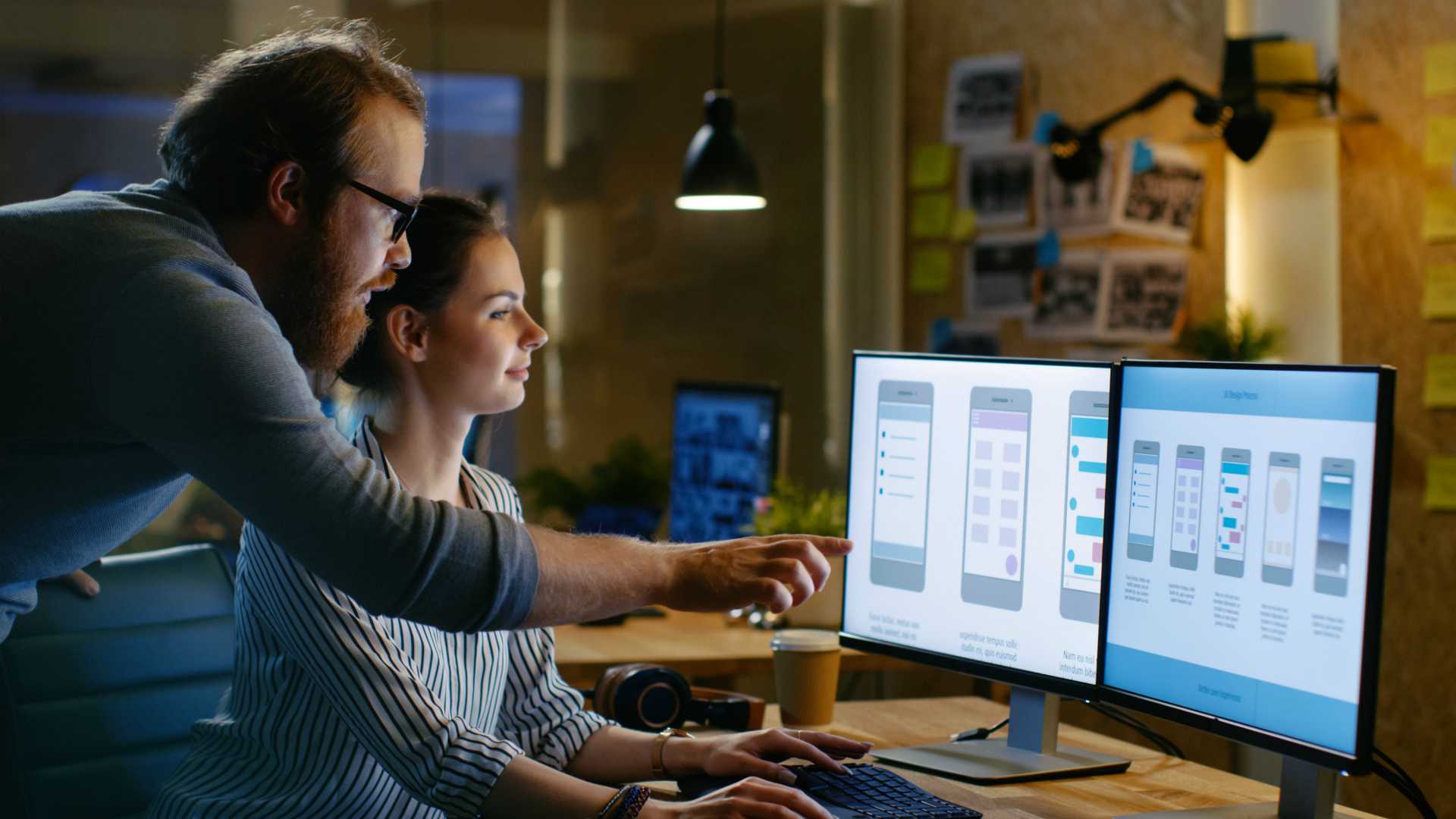 Female UX Architect Has Discussion with Male Design Engineer, They Work on Mobile Application Late at Night, She Drinks Coffee. In the Background Wall with Project Sticky Notes.