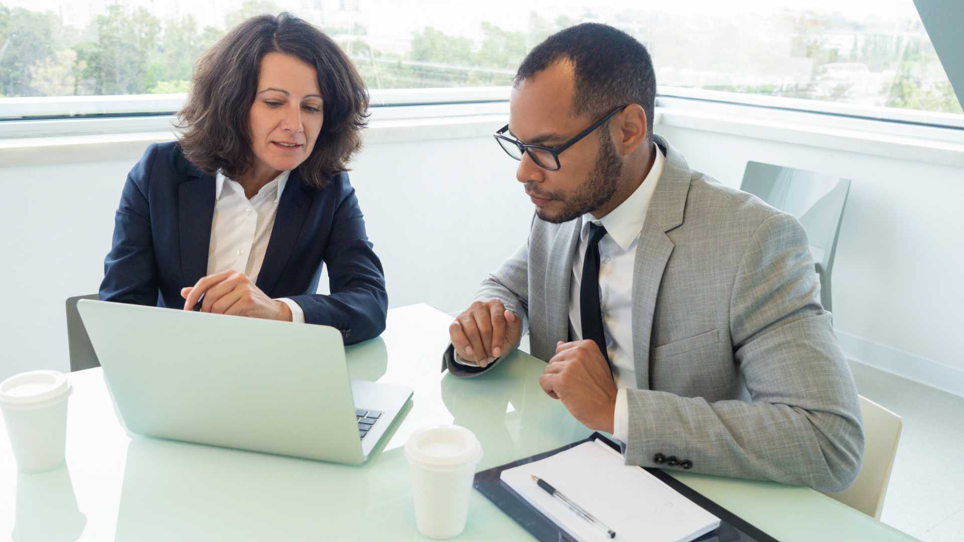 Female manager teaching male newcomer how to use corporate software. Business man and woman sitting at meeting table with open laptop and looking at screen.