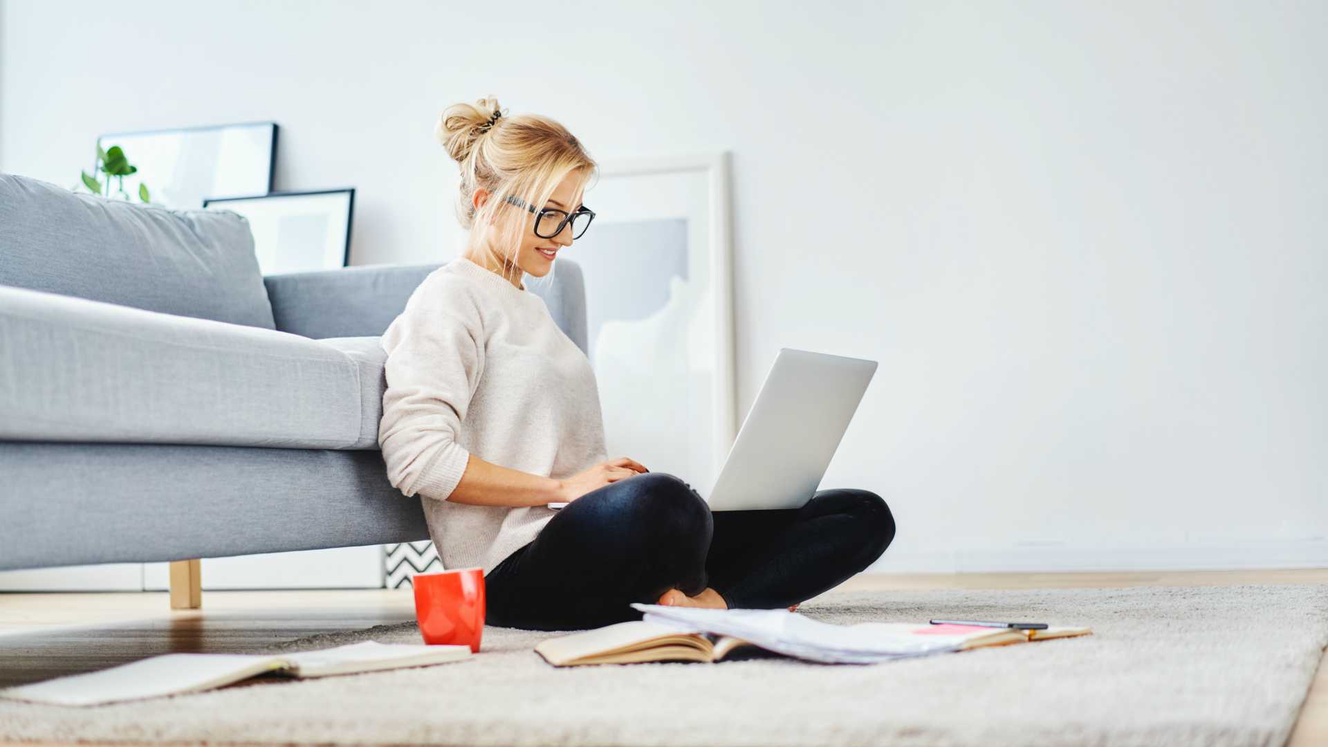 Young blonde businesswoman working remotely, sitting on her floor in front of her couch with her laptop and mug