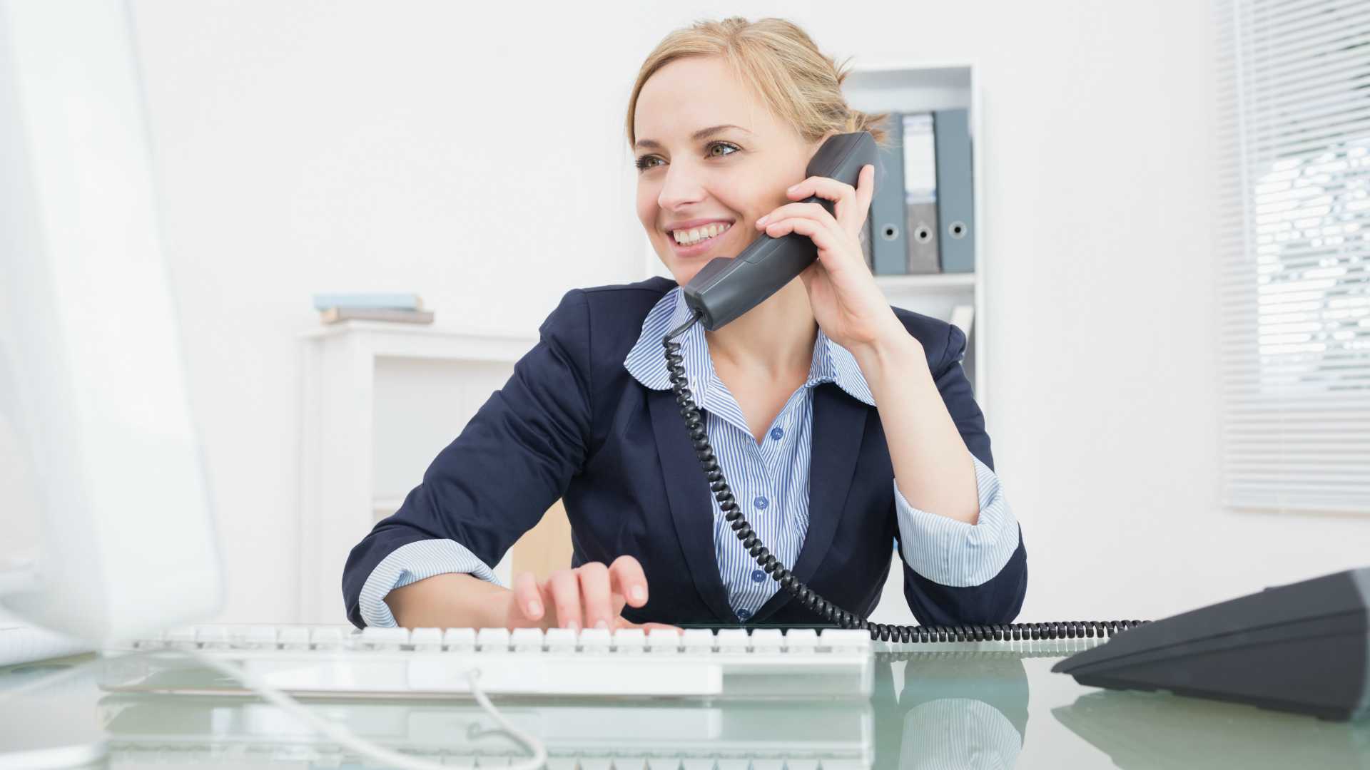 Young saleswoman working on computer and chatting on phone with client