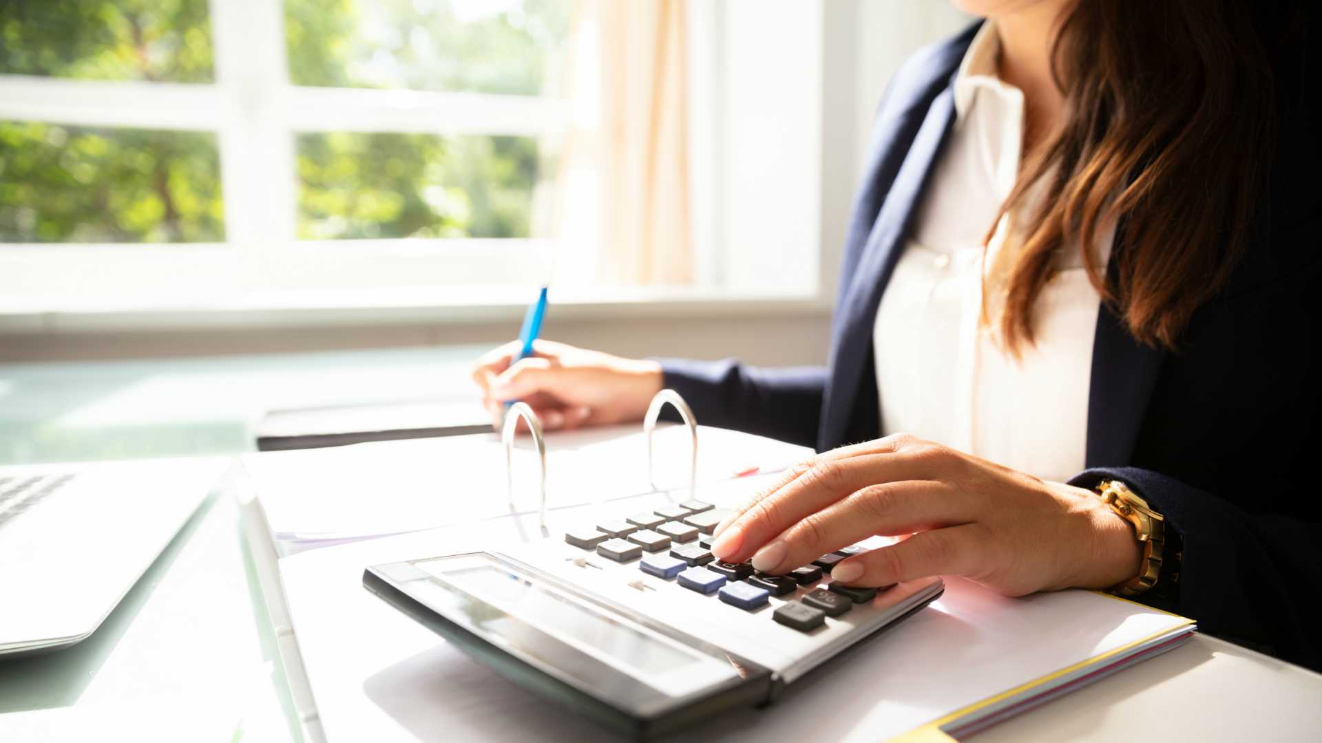Businesswoman sitting at desk, jotting notes in a binder and inputing numbers in calculator
