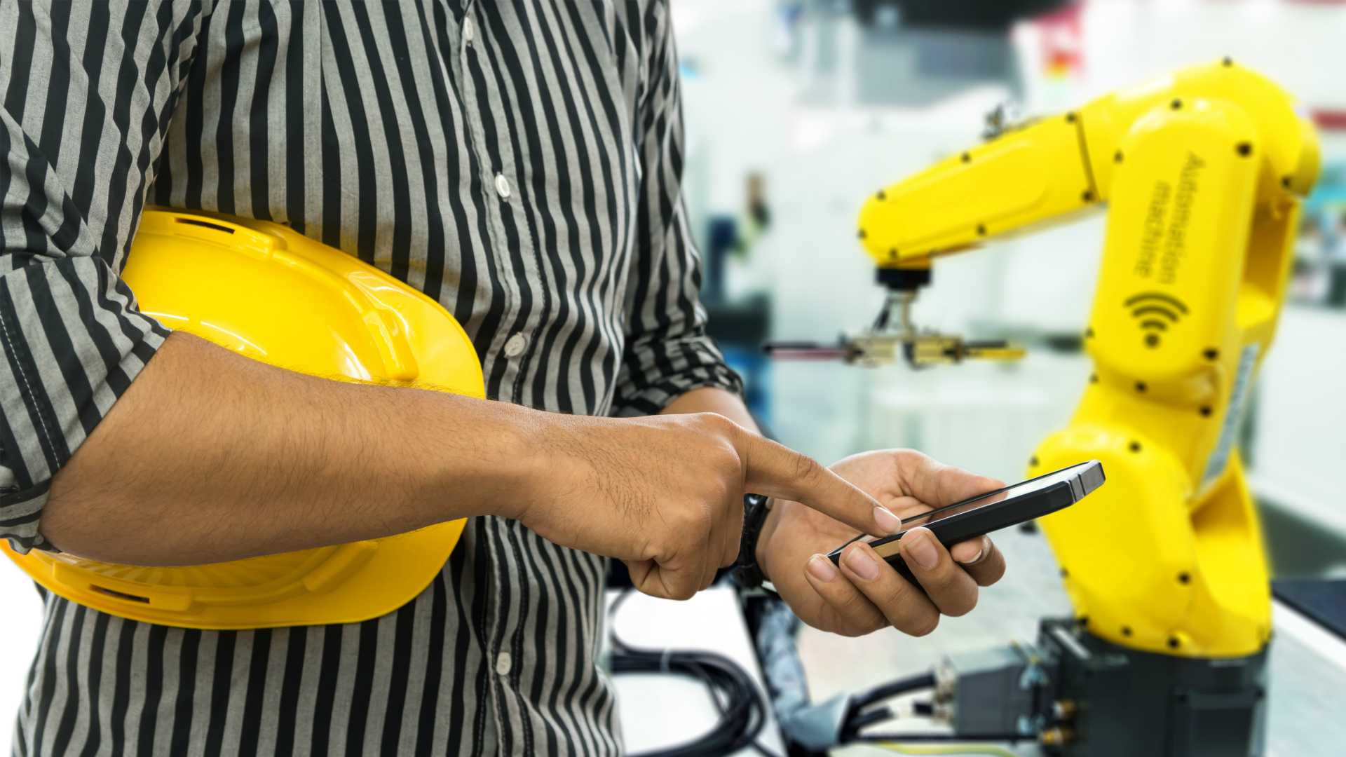 Selective focus on Engineer finger point to smart phone for control blur robot machine arm background and yellow hat in smart factory.