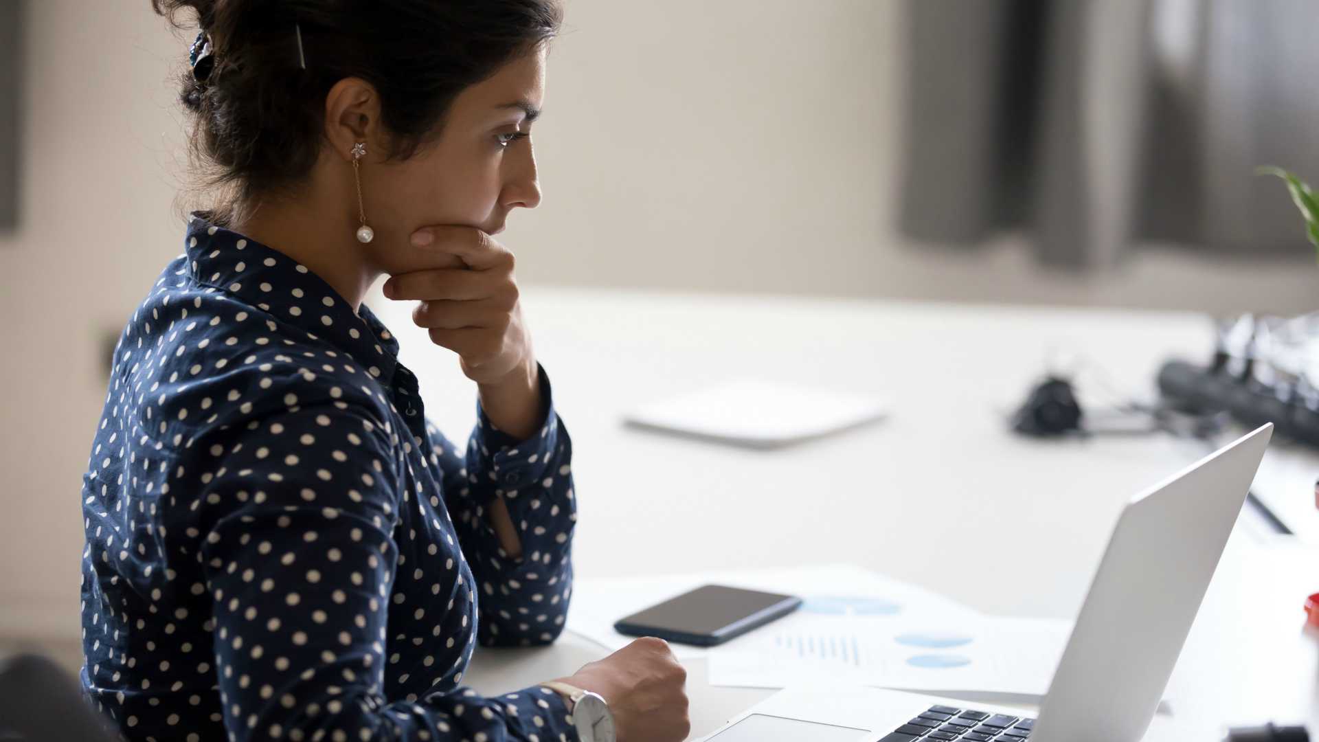 Indian girl sitting at desk near computer cogitating thinking making important decision at workplace. Concentrated serious office worker millennial woman analysing results feels doubts and feel unsure