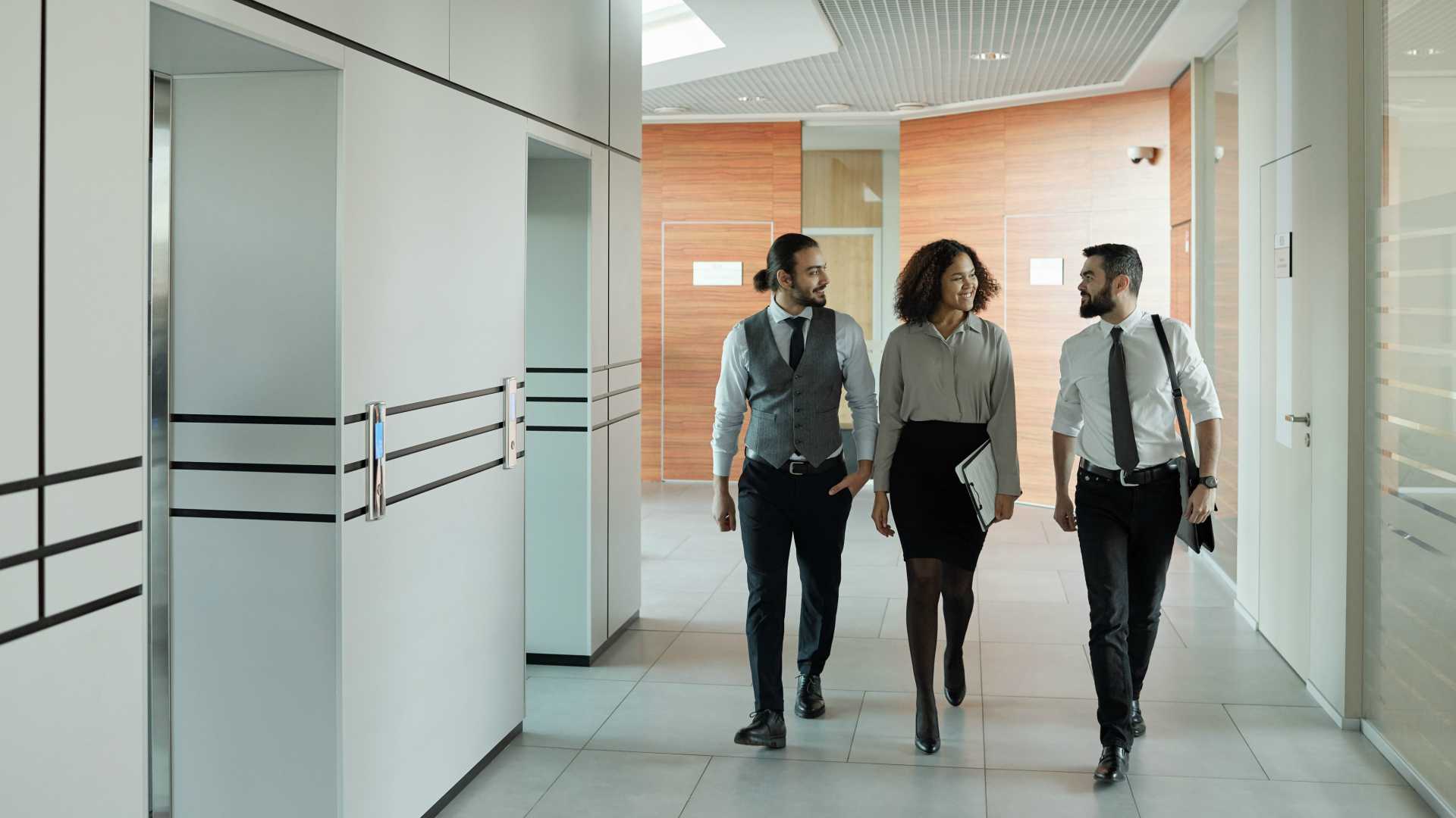Three diverse businesspeople walking down hallway in modern office building and chatting