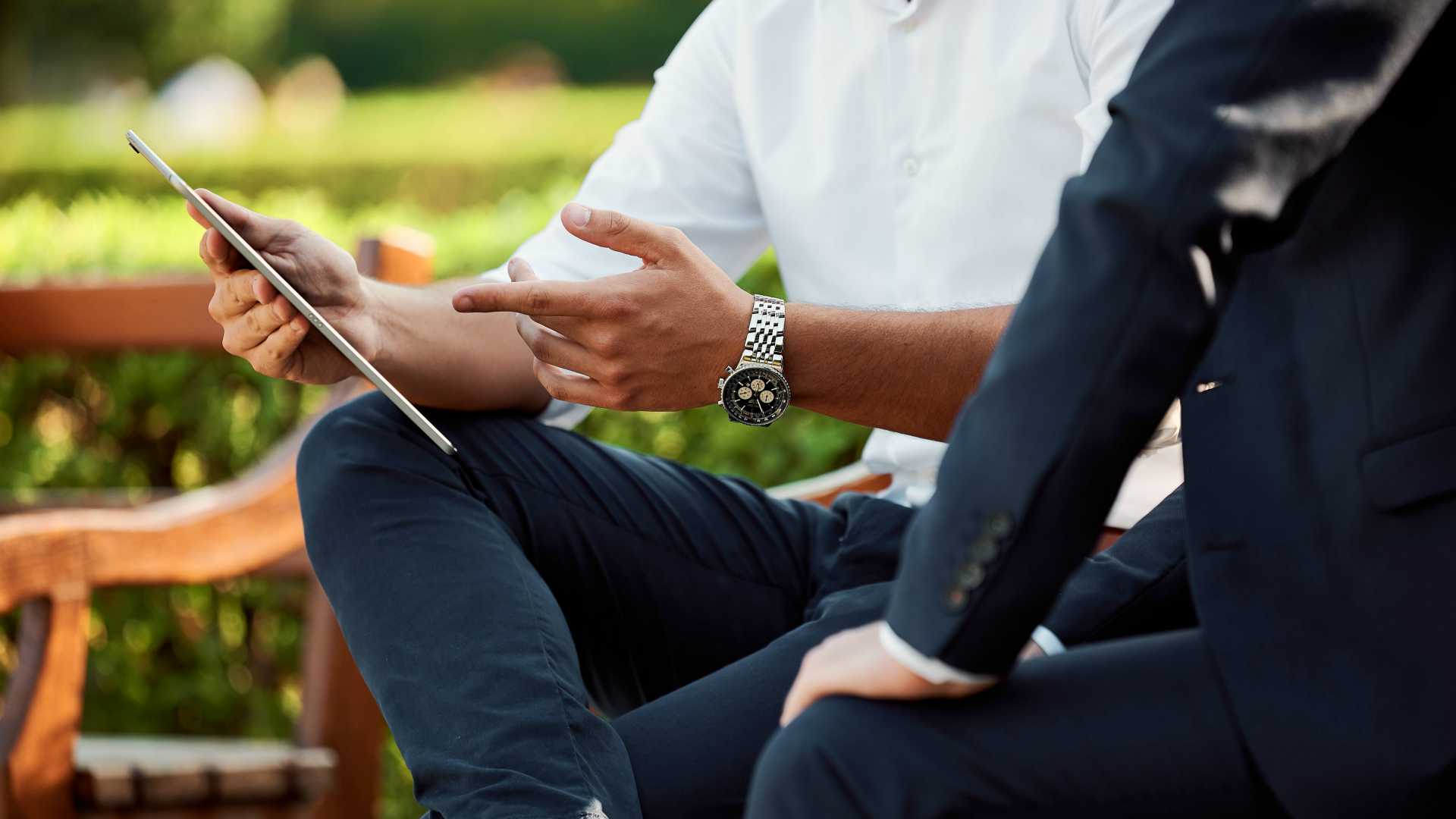 Close up shot of two businessmen sitting on bench outside. One holding a tablet and explaining business-related issue to colleague.