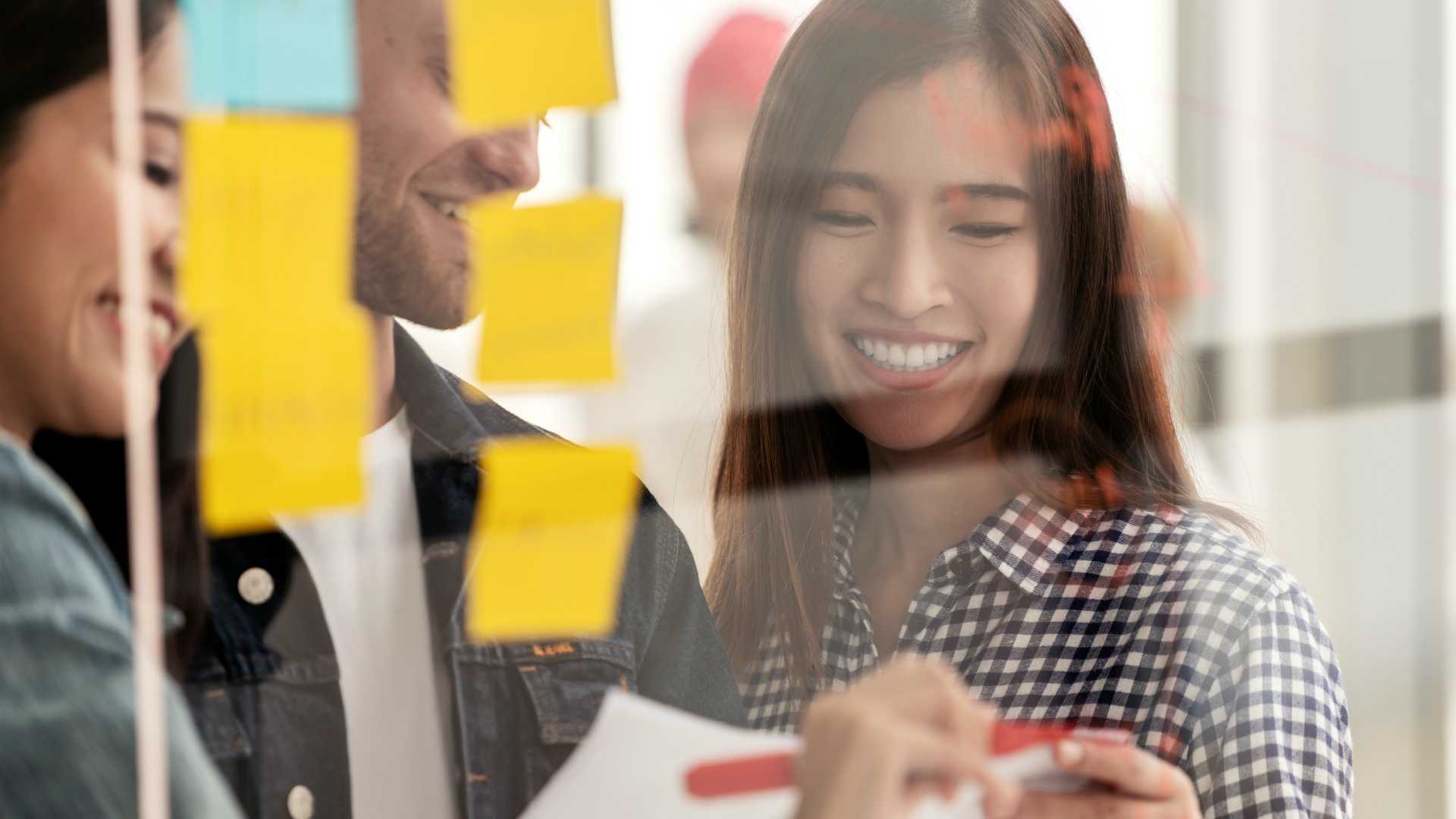 Young successful creative woman and team smile and brainstorm on project at office concentrate on paper, yellow post note and sticky note. Close up view of group sharing idea on glass wall concept.