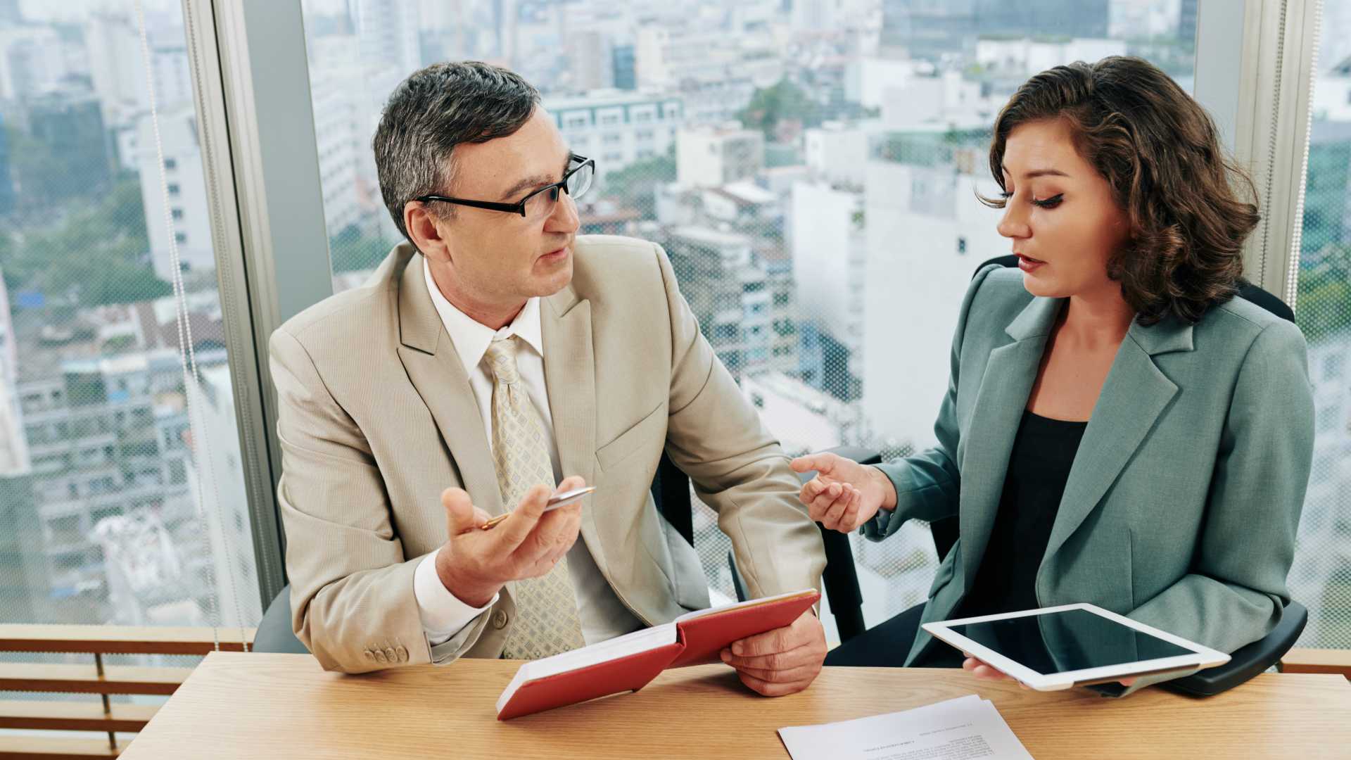 Male learning professional and female IT professional collaborating in officespace, sitting in front of window with cityscape in the background