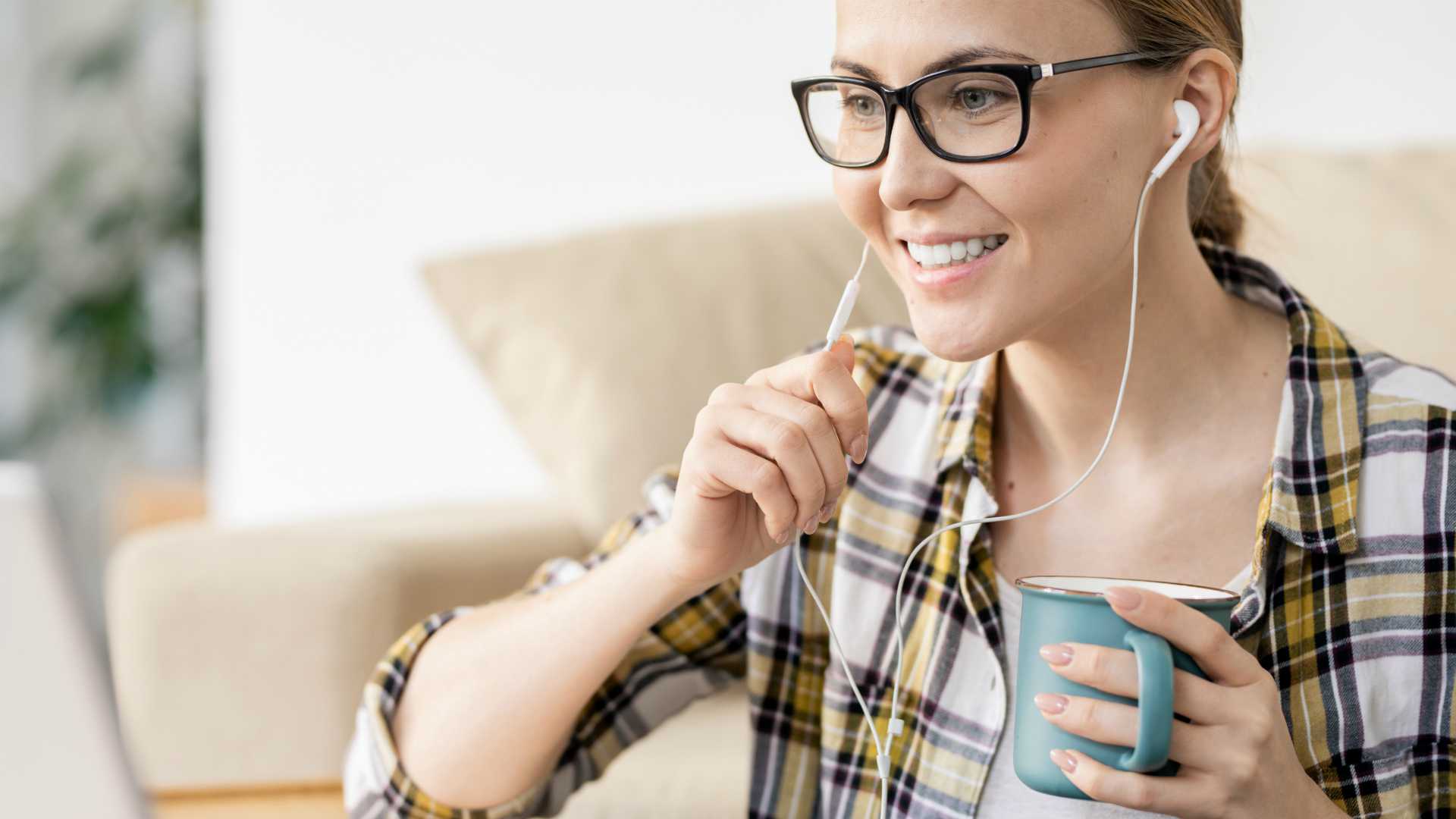 Cheerful confident attractive female remote manager in glasses drinking coffee and using laptop while answering online call using earphones