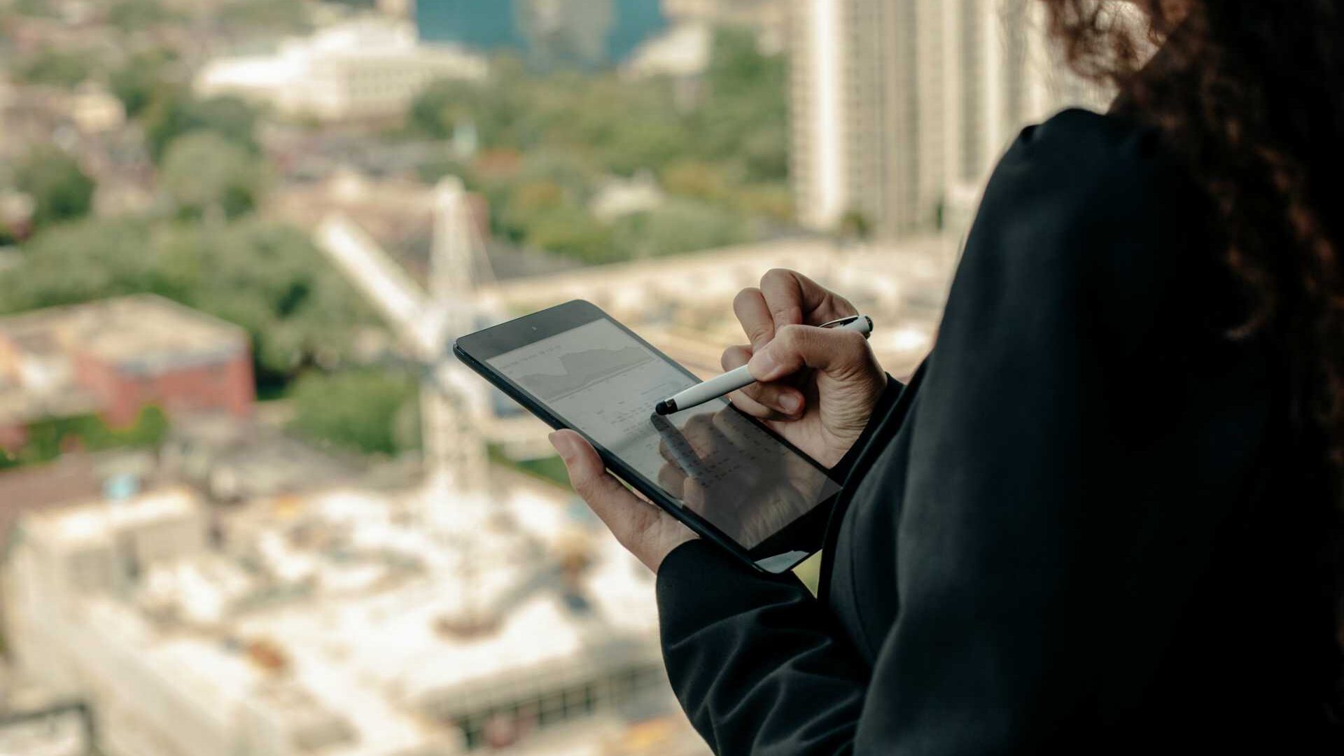 Curly-haired businesswoman holding tablet and looking out large window at cityscape below