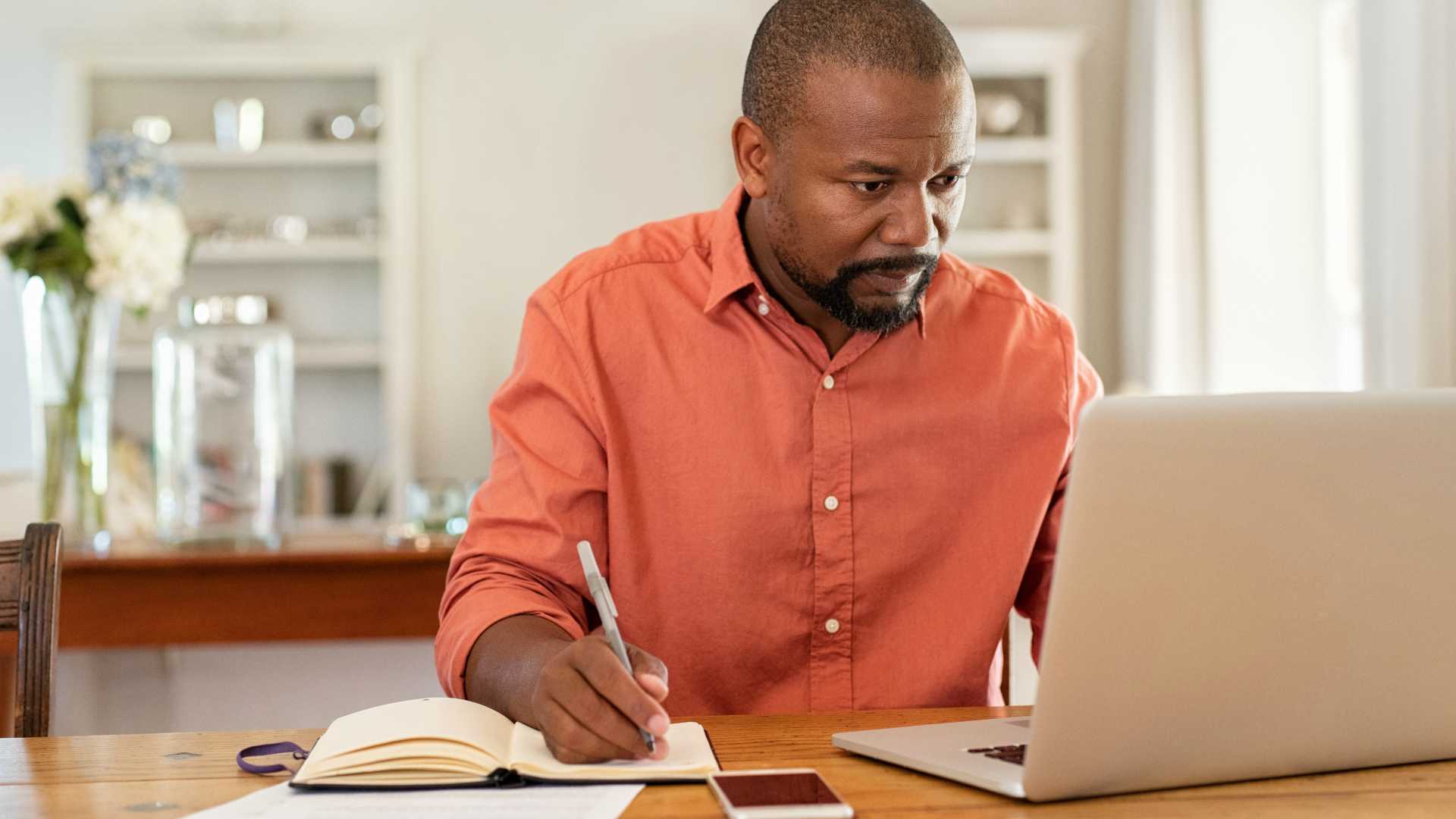 Mature man working on laptop while taking notes. Businessman working at home with computer while writing on agenda.