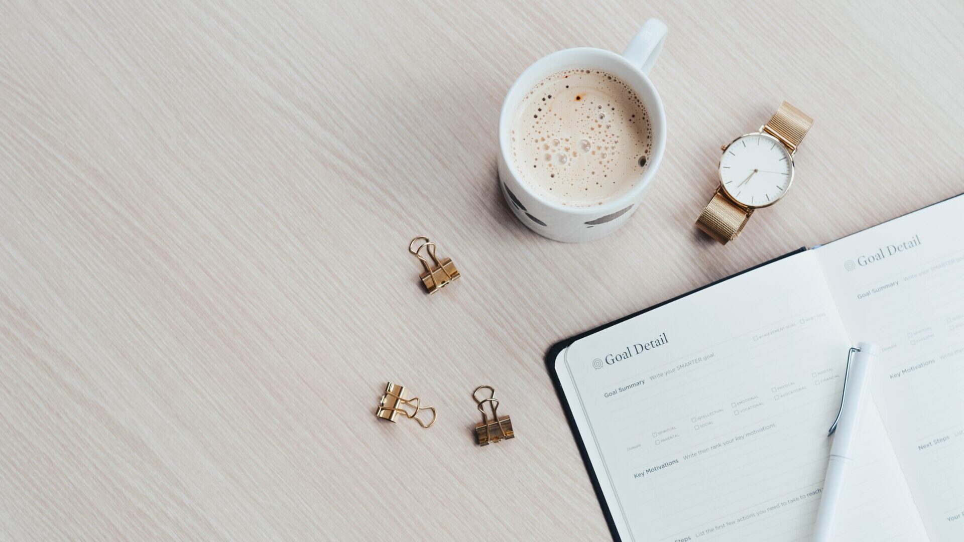 Planner on light tan desk with cup of coffee, watch and paper binders scattered around