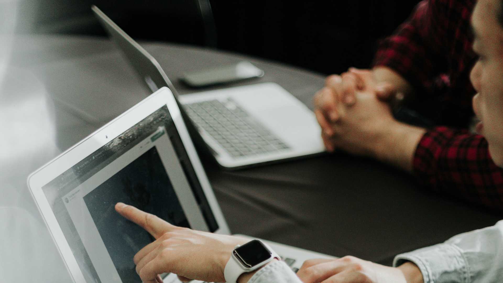 Two businesspeople working side-by-side. Businssperson in foreground pointing at laptop screen; businessperson in background sitting with hands crossed and listening.