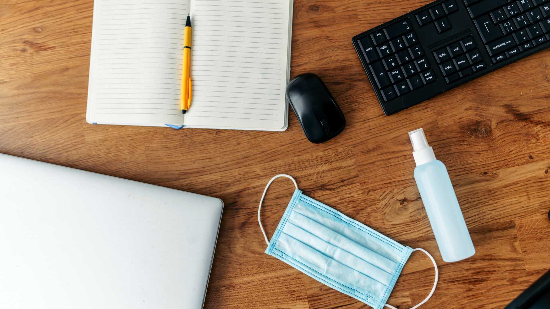 Aerial view of wooden desk with face mask, hand sanitizer, keyboard and mouse, and open notebook