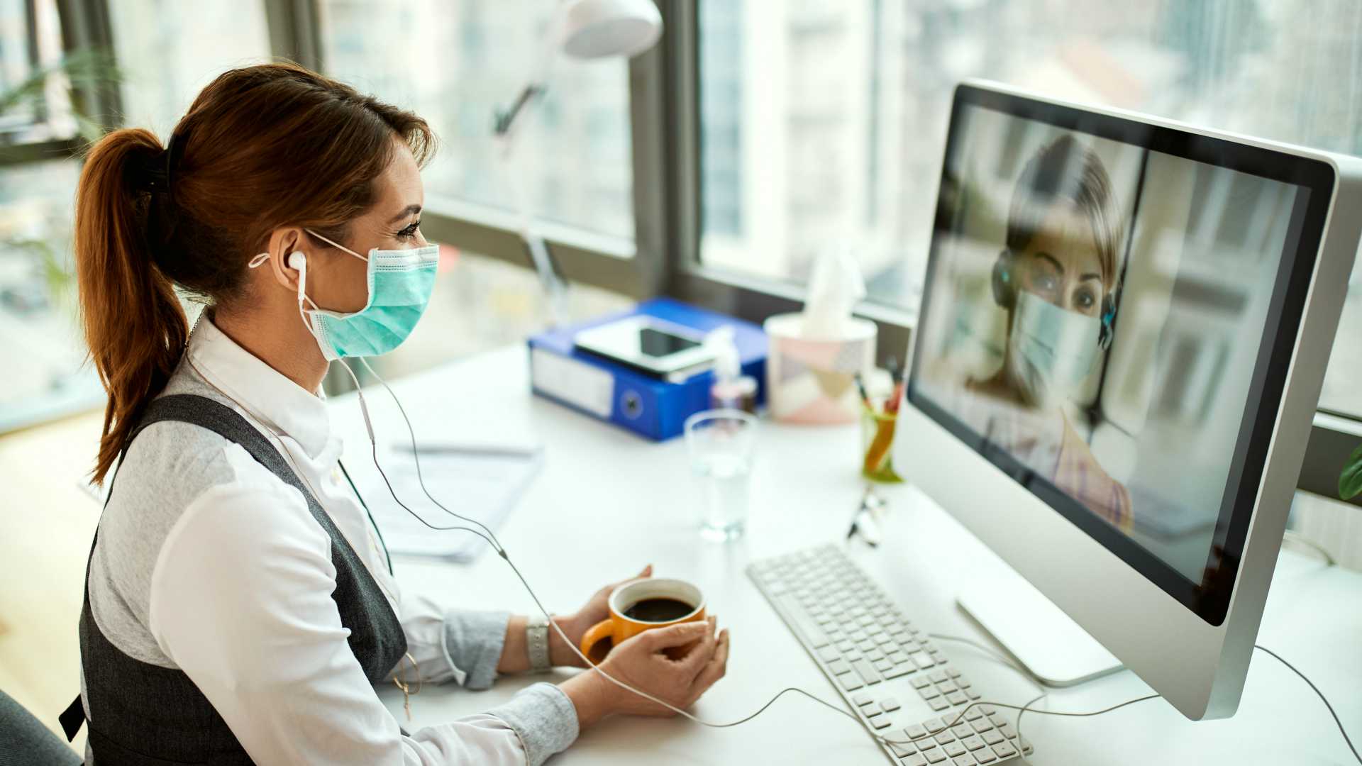 Smiling businesswoman with face mask using computer and making video call while having coffee break in the office.