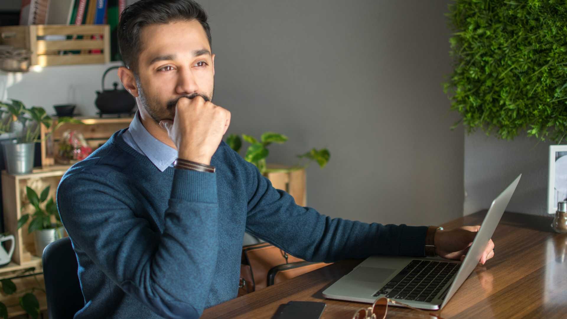 Pensive young bearded businessman holding hand on jaw and looking away while working on laptop at office.